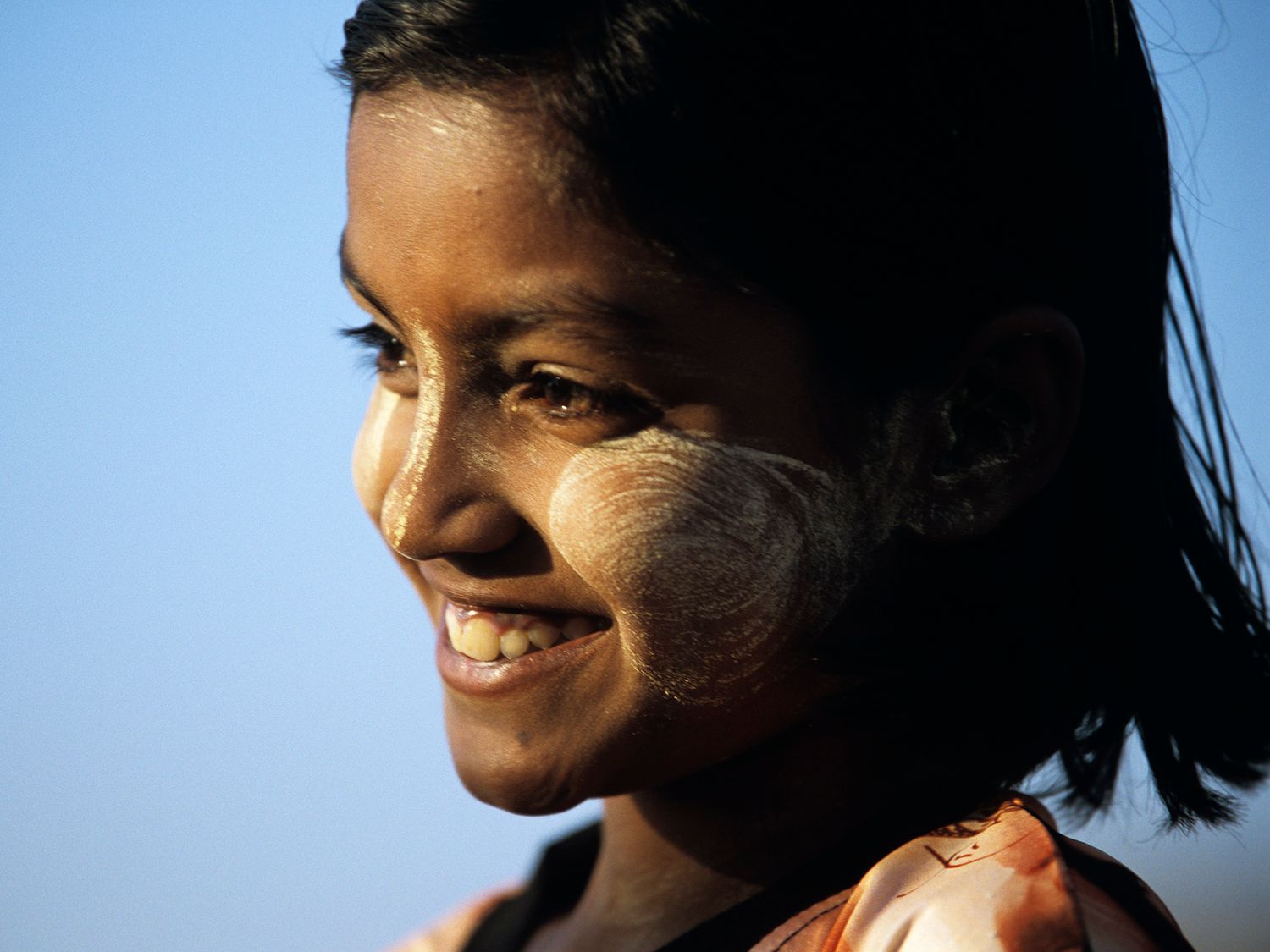 Smiling young girl with traditional thanaka paste on her cheeks against a blue sky background. Burma, Myanmar