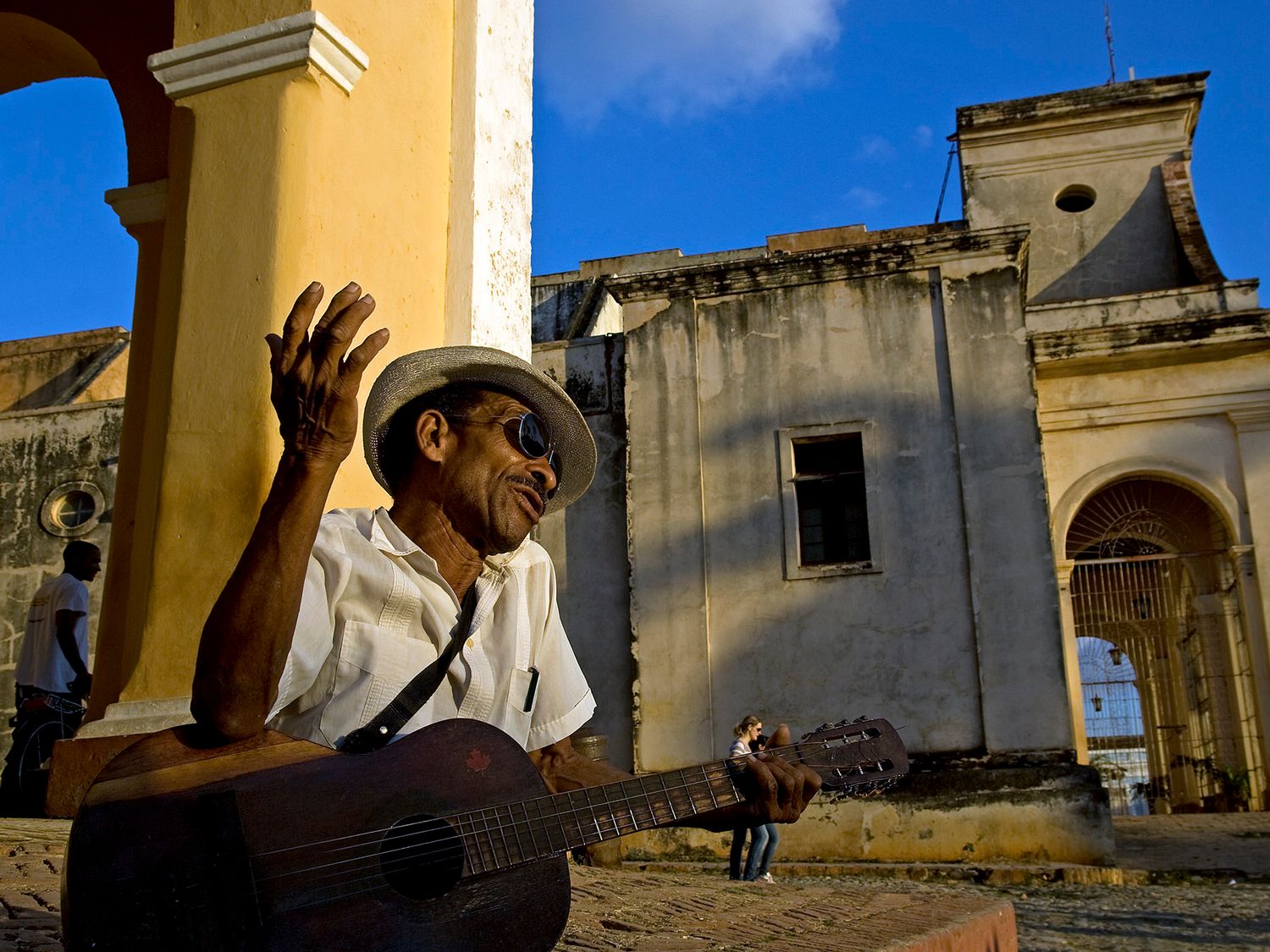 Elderly Cuban musician playing guitar outdoors near colonial architecture in Trinidad, Cuba, wearing a straw hat.
