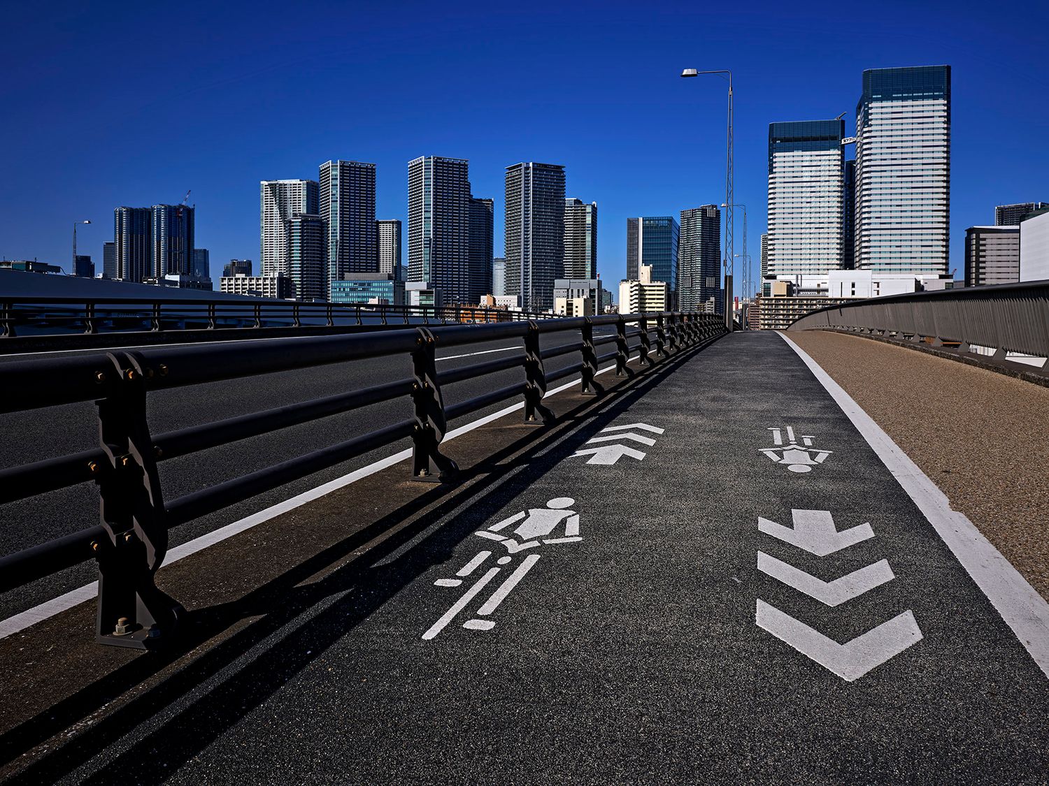 A pedestrian and cycling path on a bridge with white arrow markings leads toward a modern city skyline under a clear blue sky.