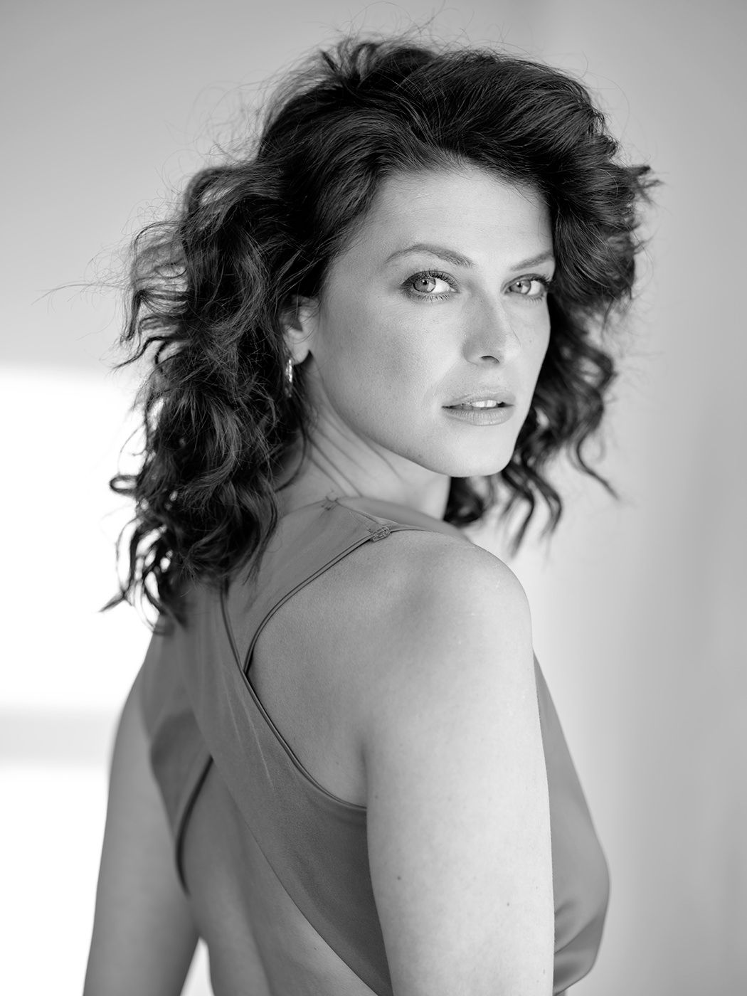 Black and white portrait of a woman with curly hair in a slip dress, glancing back over her shoulder.