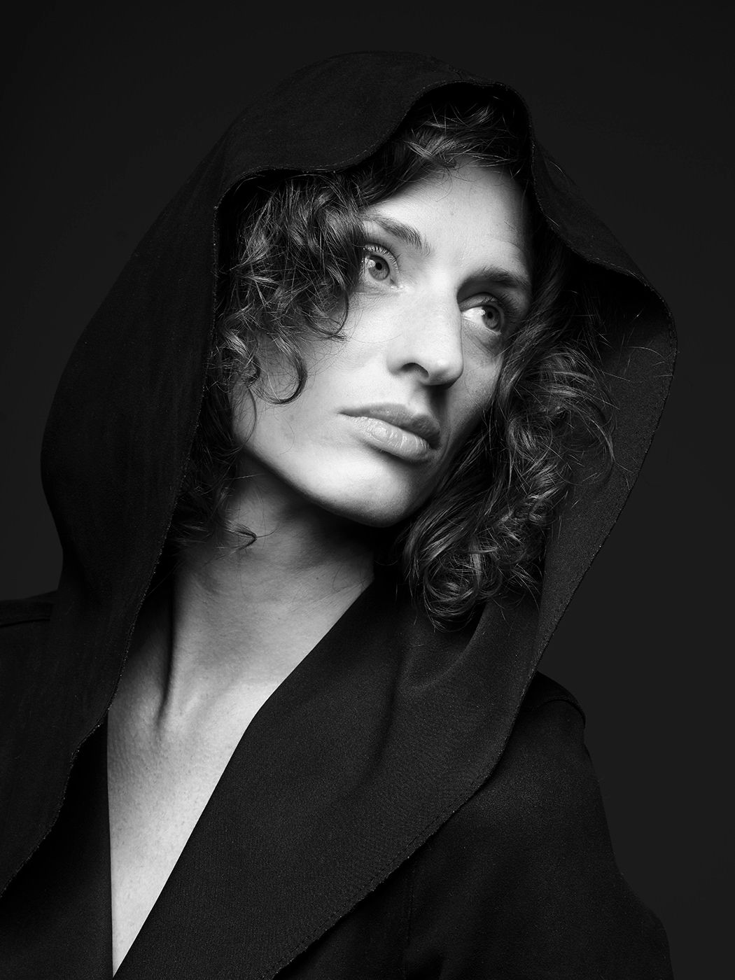 Dramatic black and white portrait of a woman in a hooded black cloak gazing upward.