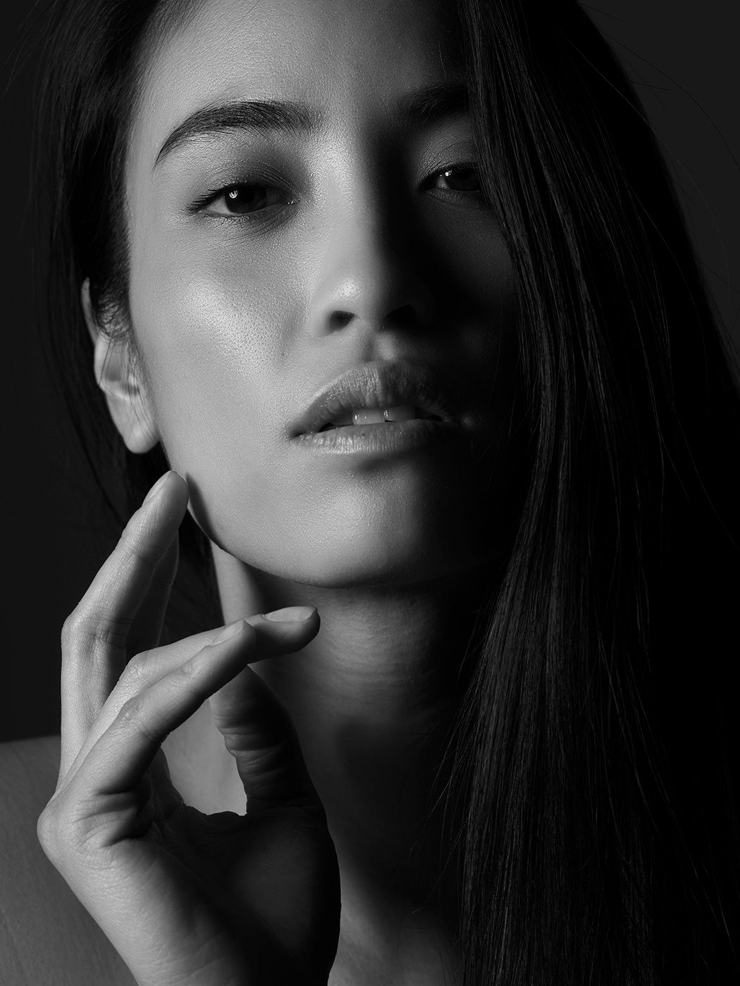 Black and white portrait of a young Asian woman with long hair, gently touching her chin, dramatic lighting.