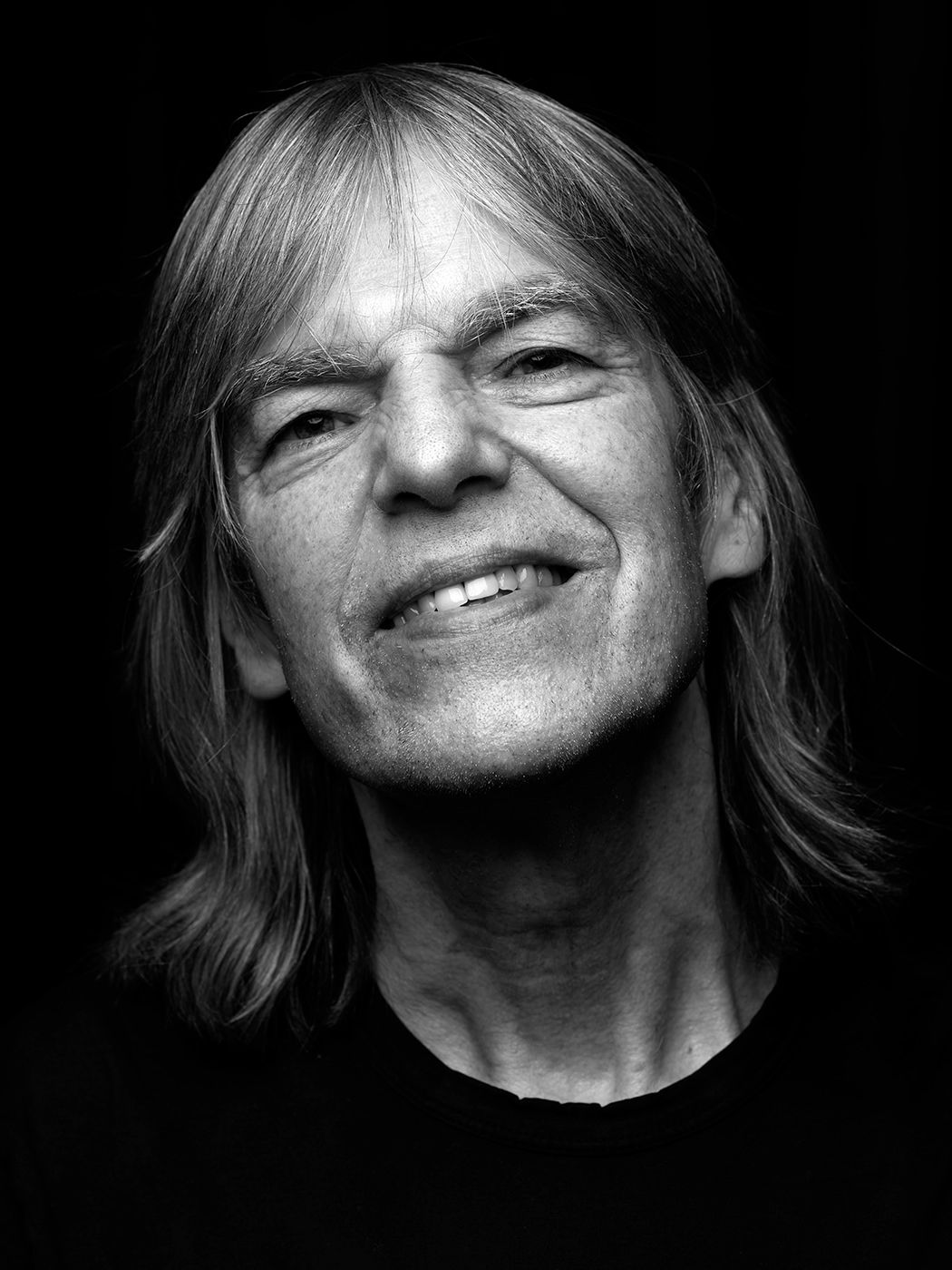 Black and white portrait of a smiling middle-aged man with long hair wearing a dark shirt. Mike Stern