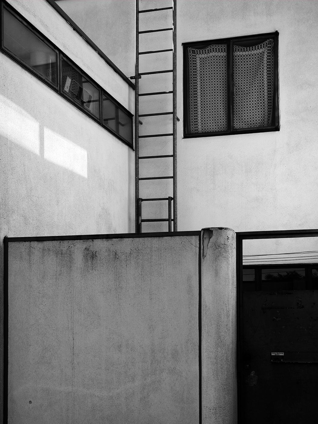 Black and white photo of a building exterior with a metal ladder, textured walls, and a window with decorative grille.