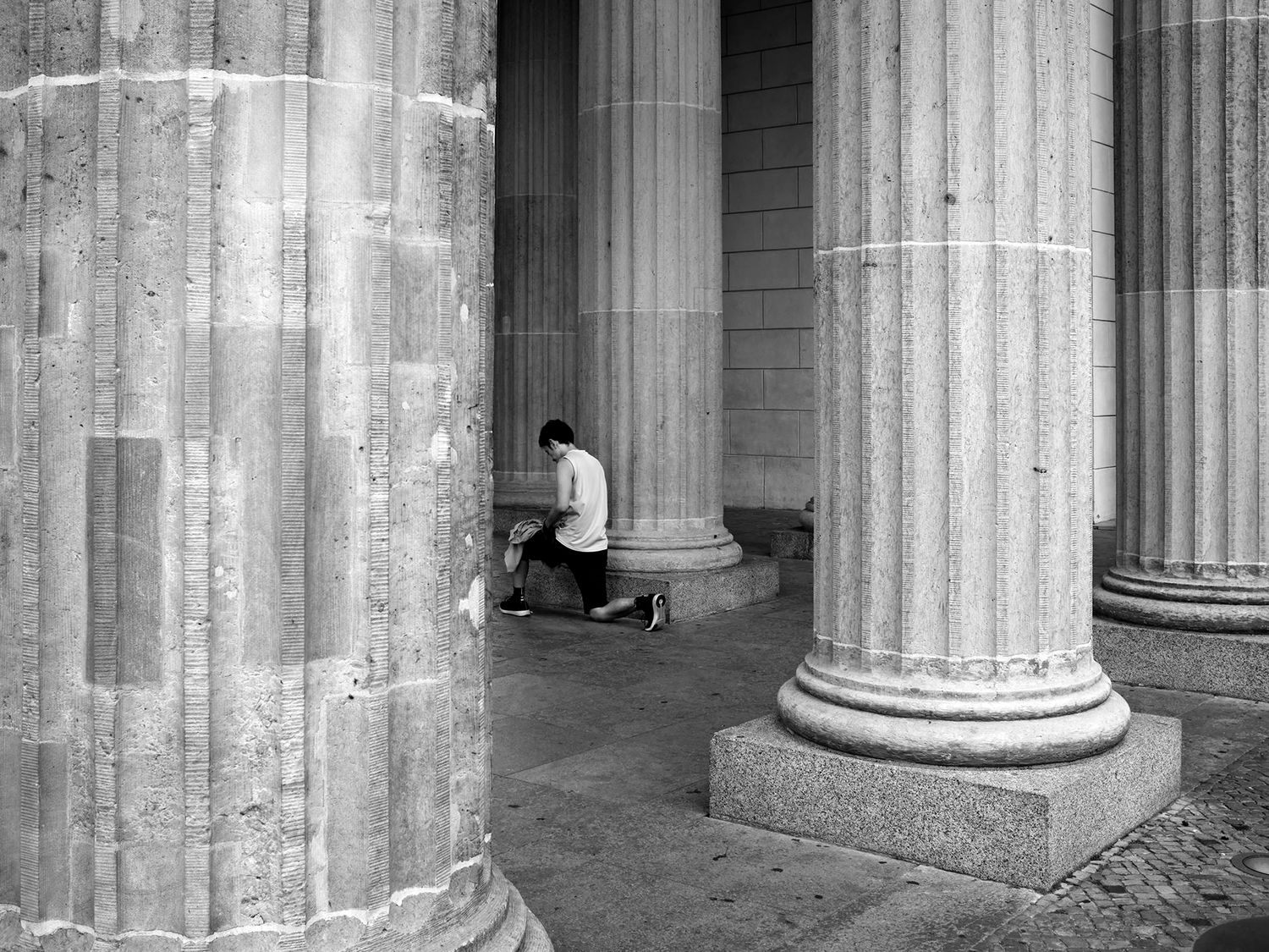 Black and white photo of a person kneeling between grand stone columns of a classical building.