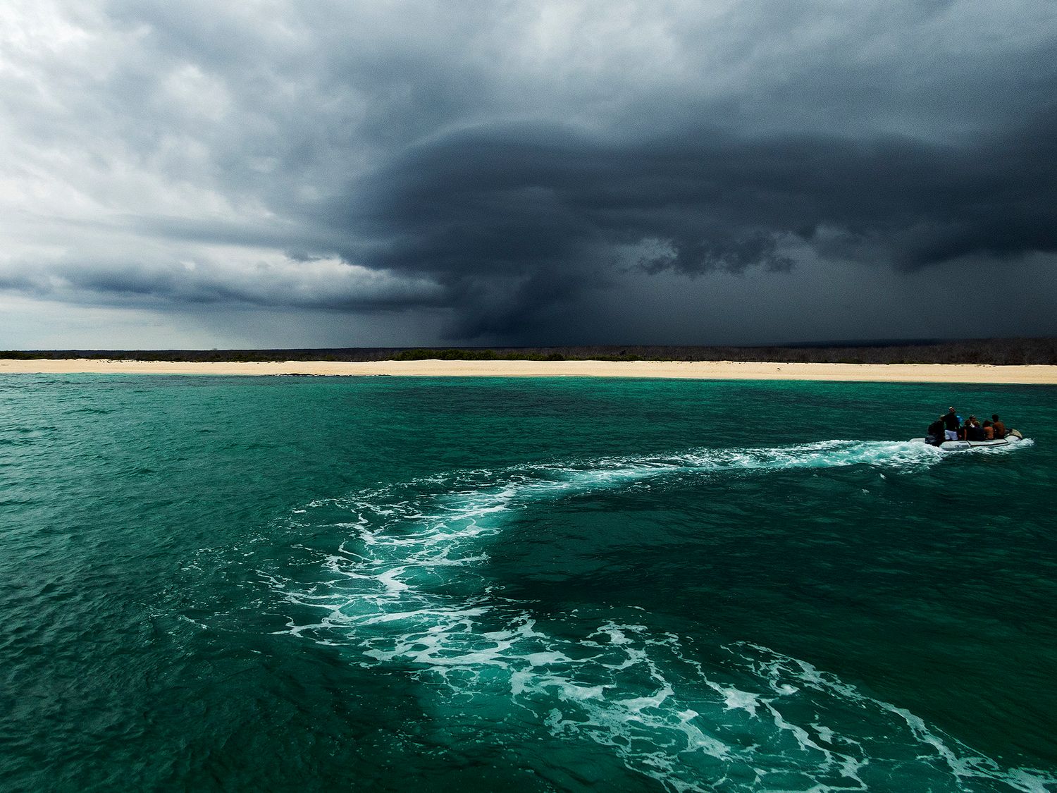 A small boat speeds across turquoise waters toward a dramatic storm cloud over a sandy beach shoreline.