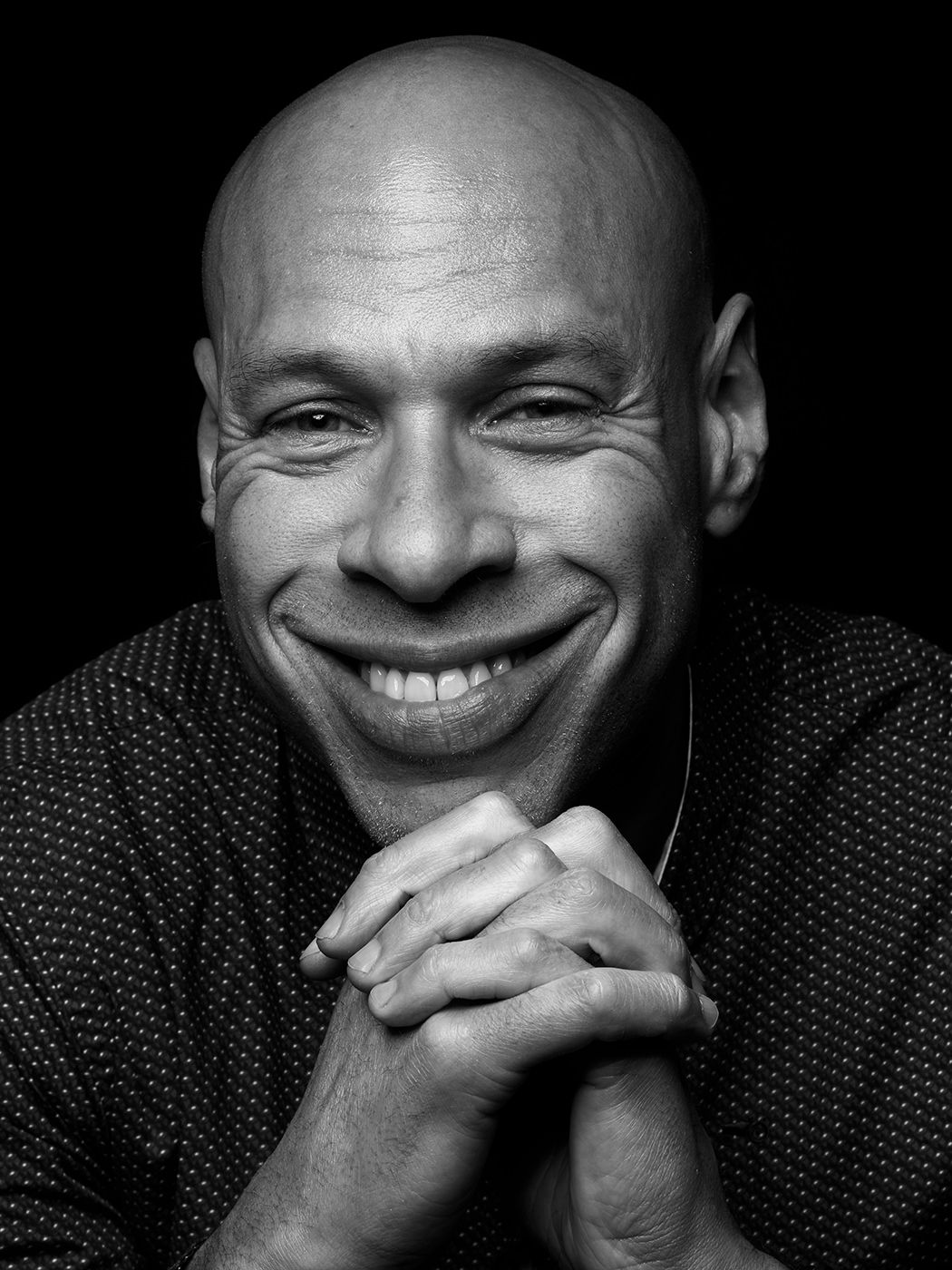 Black and white portrait of a smiling bald man with clasped hands, studio photography. Joshua Redman