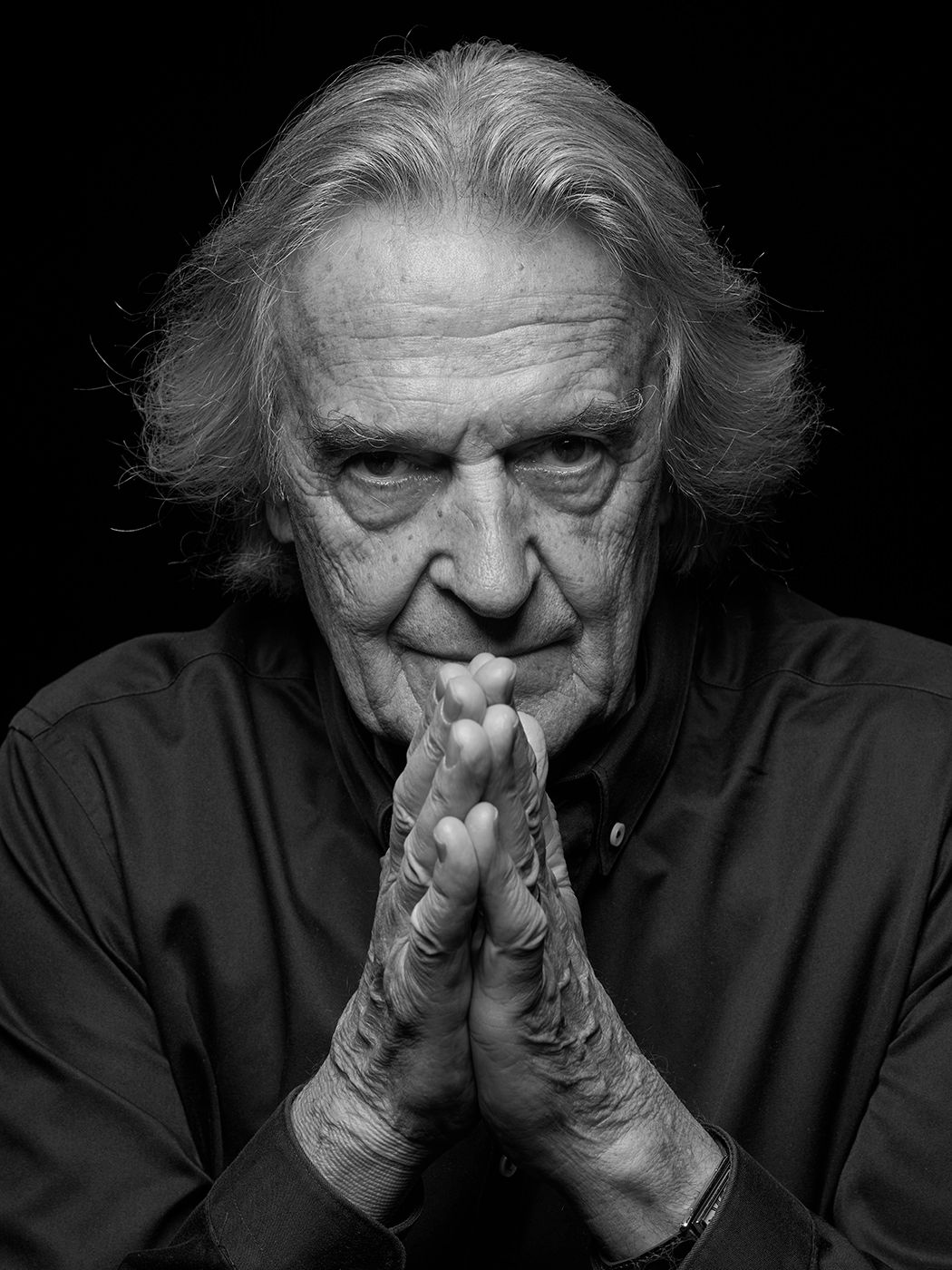 Black and white portrait of an elderly man with long gray hair, hands clasped in prayer pose, against dark background. John McLaughlin