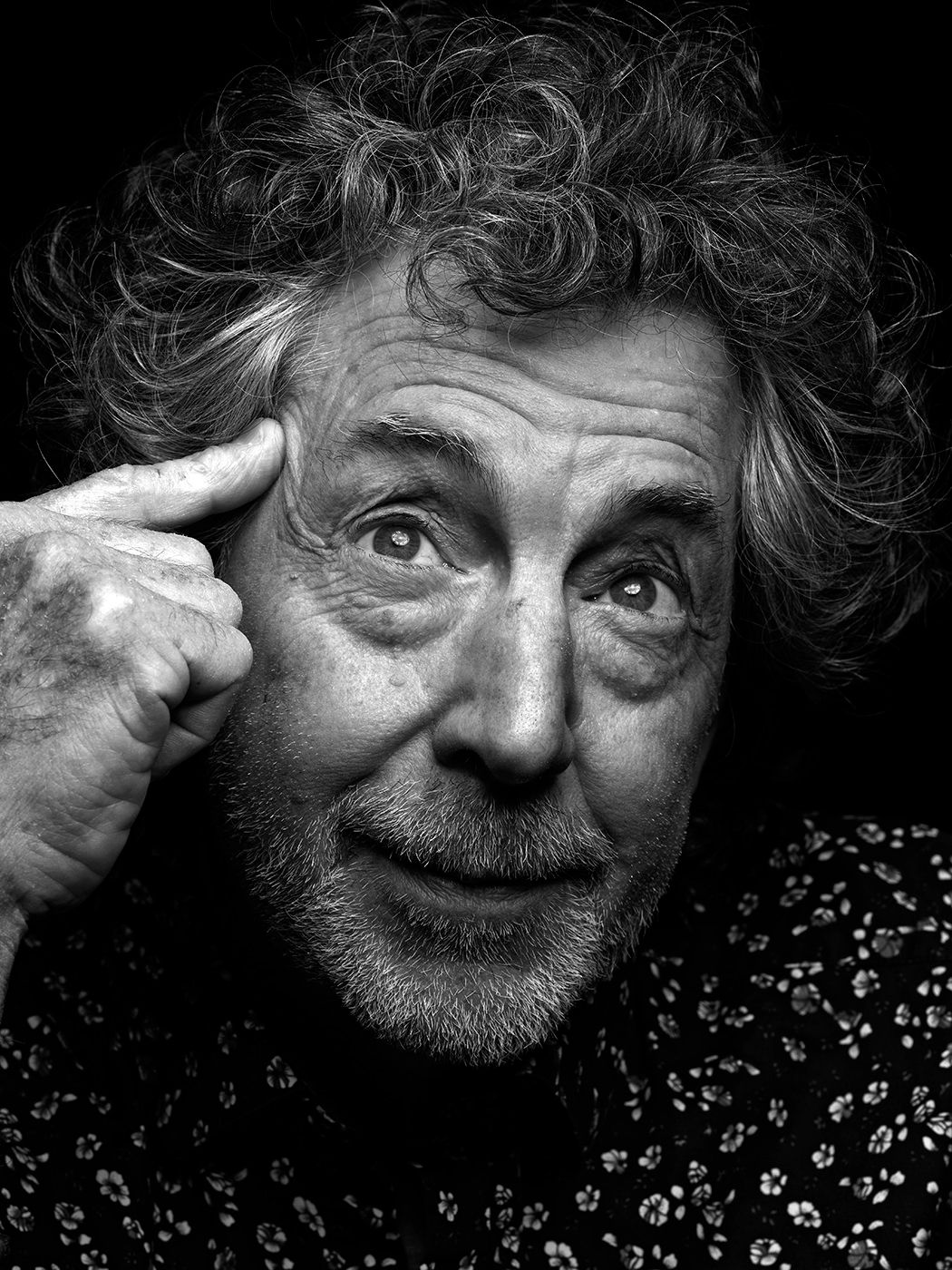 Black and white portrait of a curly-haired man pointing to his temple, smiling expressively. Simon Phillips, musician, drummer.