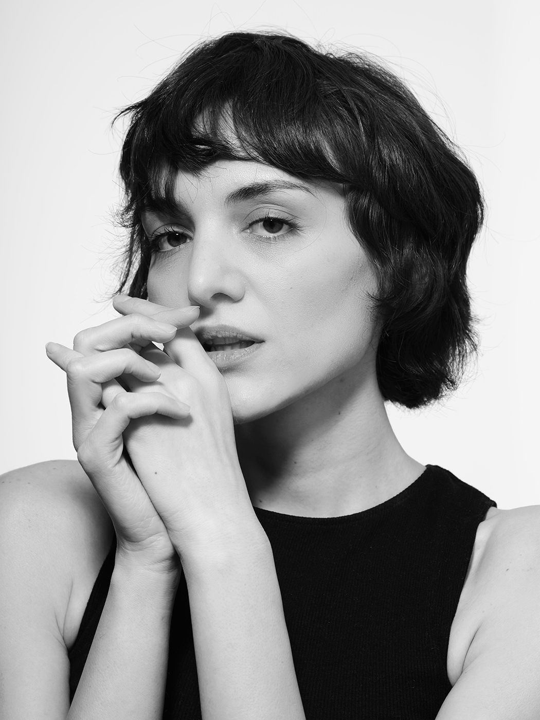 Black and white portrait of a young woman with short dark hair and bangs, hands clasped near her face.