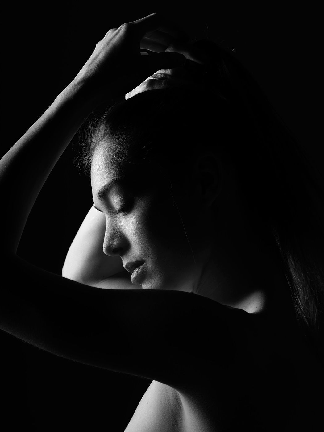 Black and white portrait of a young woman with eyes closed, hands raised to her head, dramatic low-key lighting.