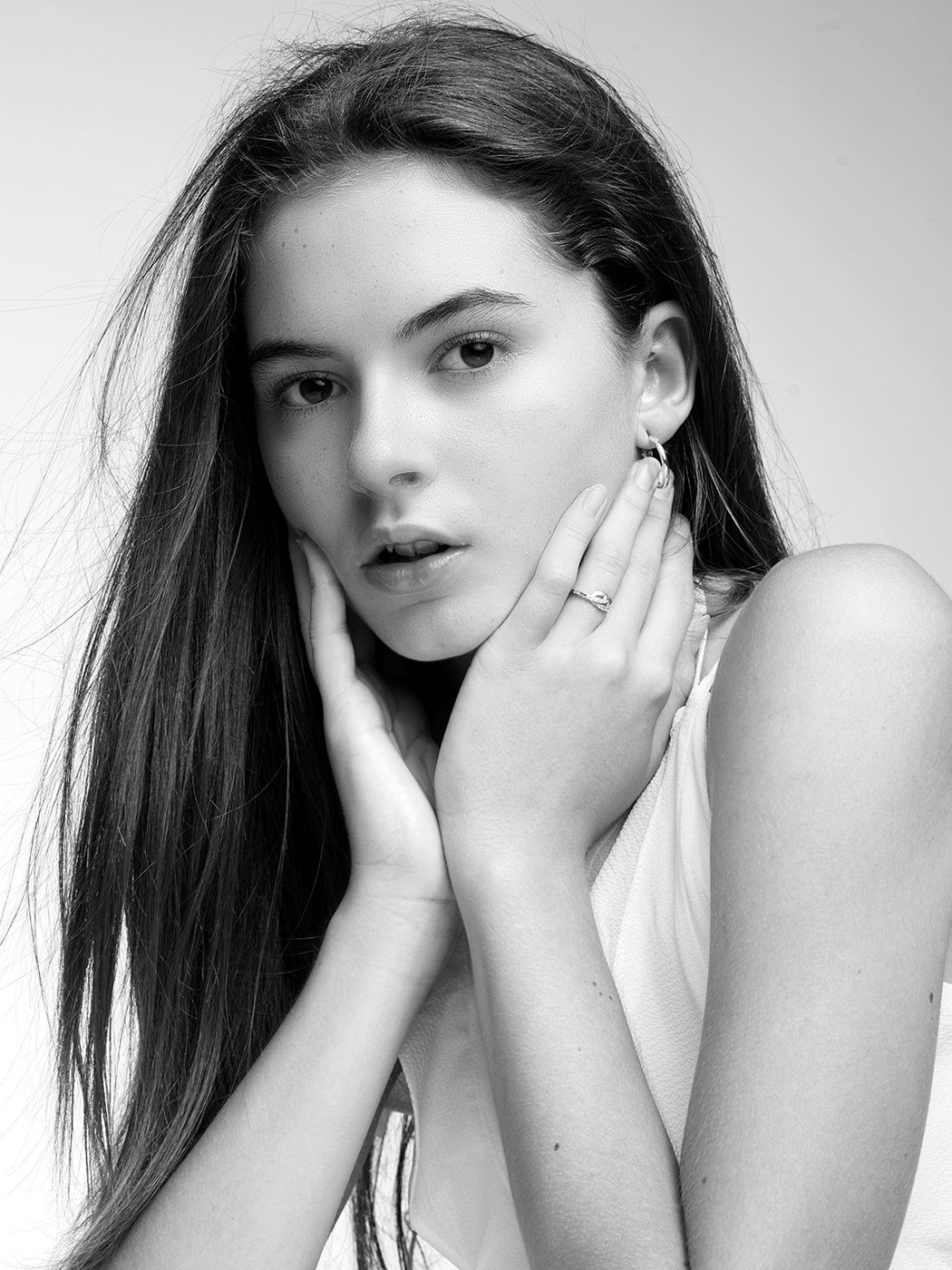 Black and white portrait of a young woman with long hair, hands framing her face, wearing a delicate ring.