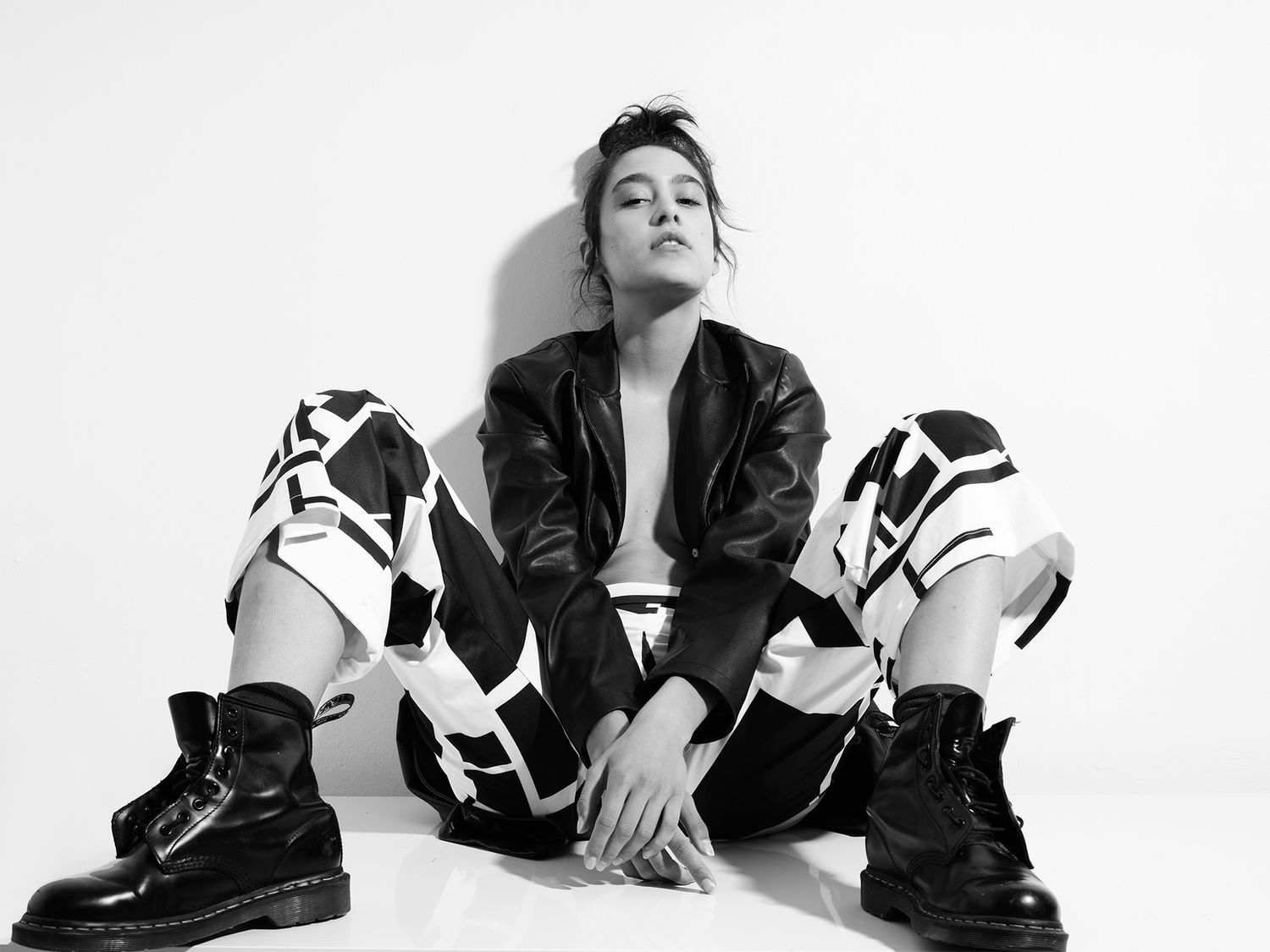 Black and white photo of a young woman in leather jacket and patterned pants sitting on floor wearing combat boots.