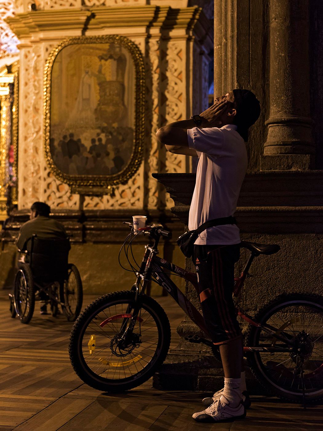 Man with bicycle stands near ornate church facade at night, while another person sits in background.