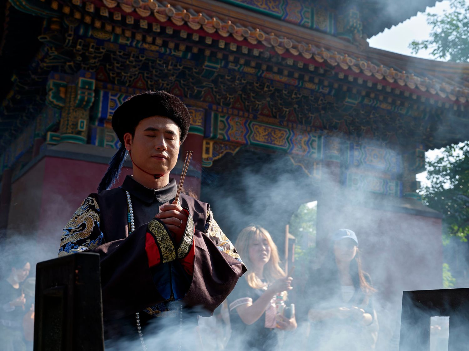 Man in traditional Chinese ceremonial robes stands amid incense smoke before a colorful temple gate.