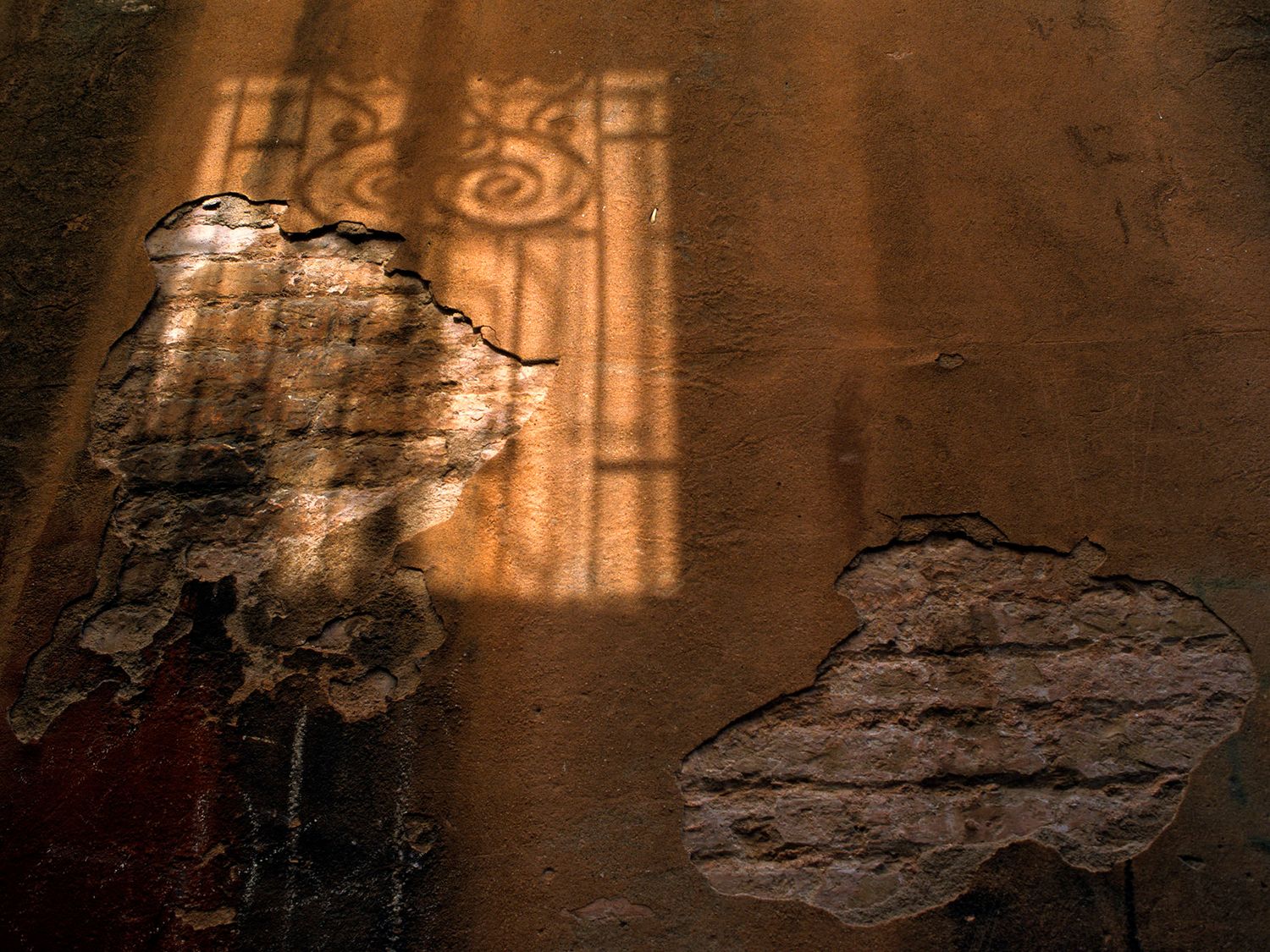 Sunlight casts ornate window shadow on aged, cracked stucco wall with exposed brick patches in warm amber tones.