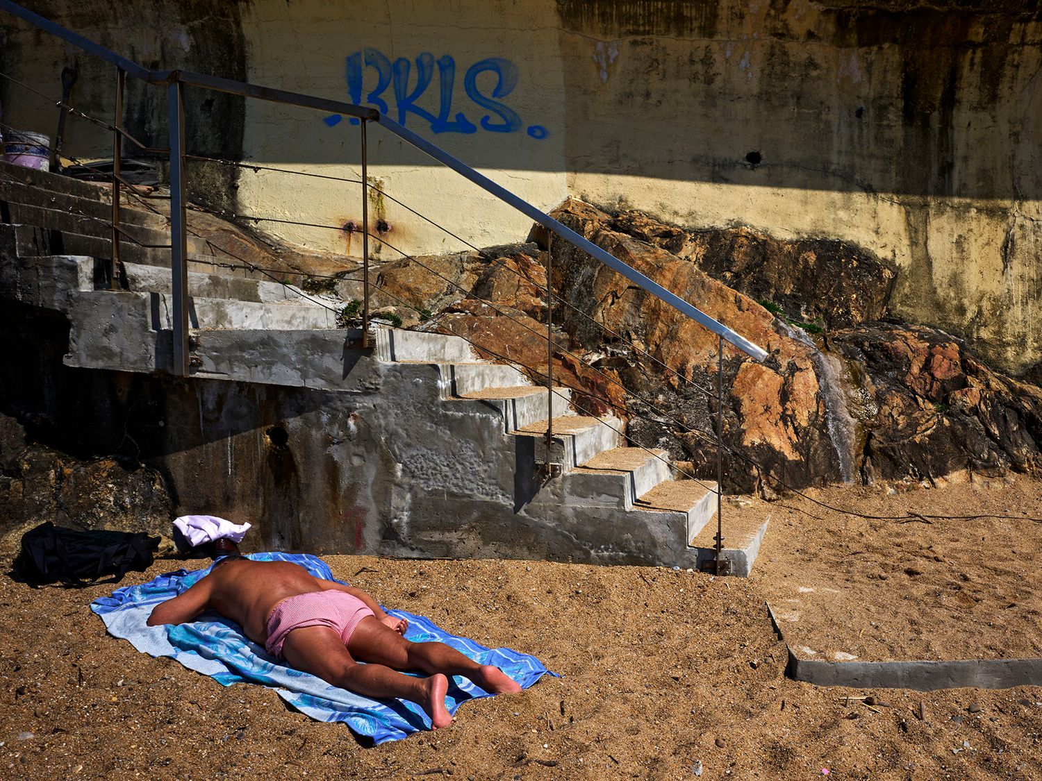 Person sunbathing on a blue towel on sandy ground near graffiti-covered concrete steps outdoors.