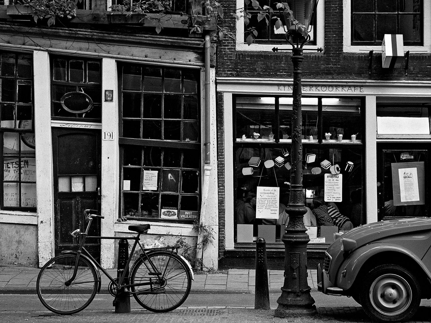Black and white street scene with a bicycle, lamppost, and vintage car outside a quaint European shop.
