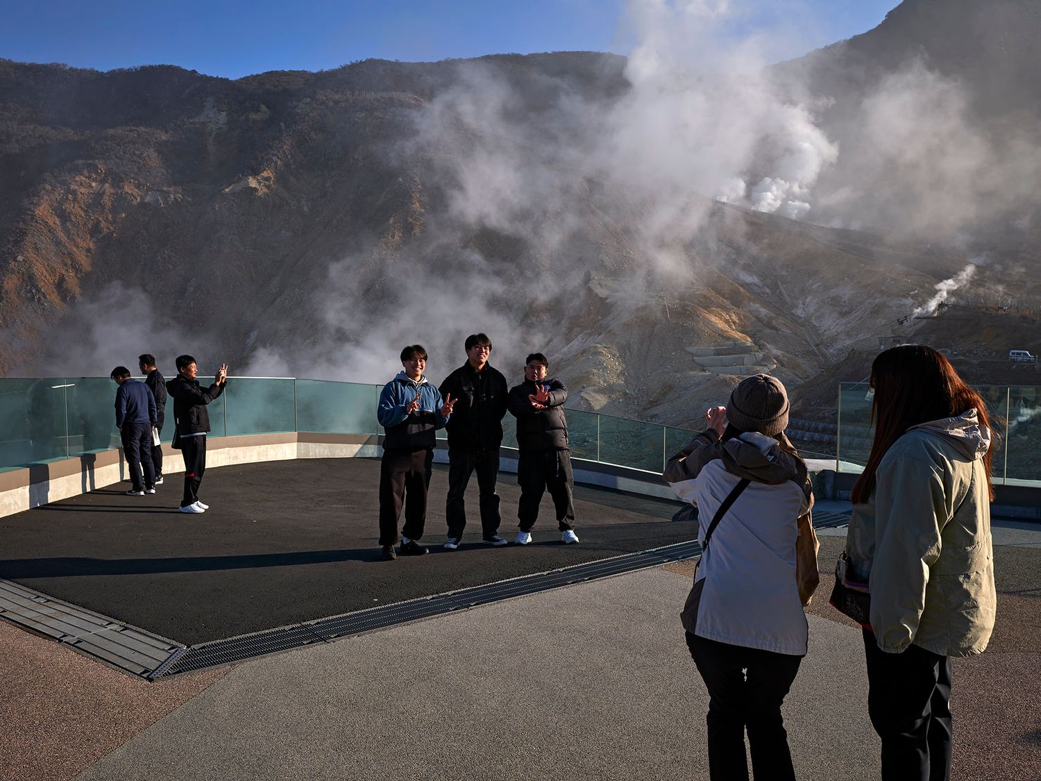 Tourists photographing friends on volcanic observation deck with steaming geothermal vents and mountain backdrop.