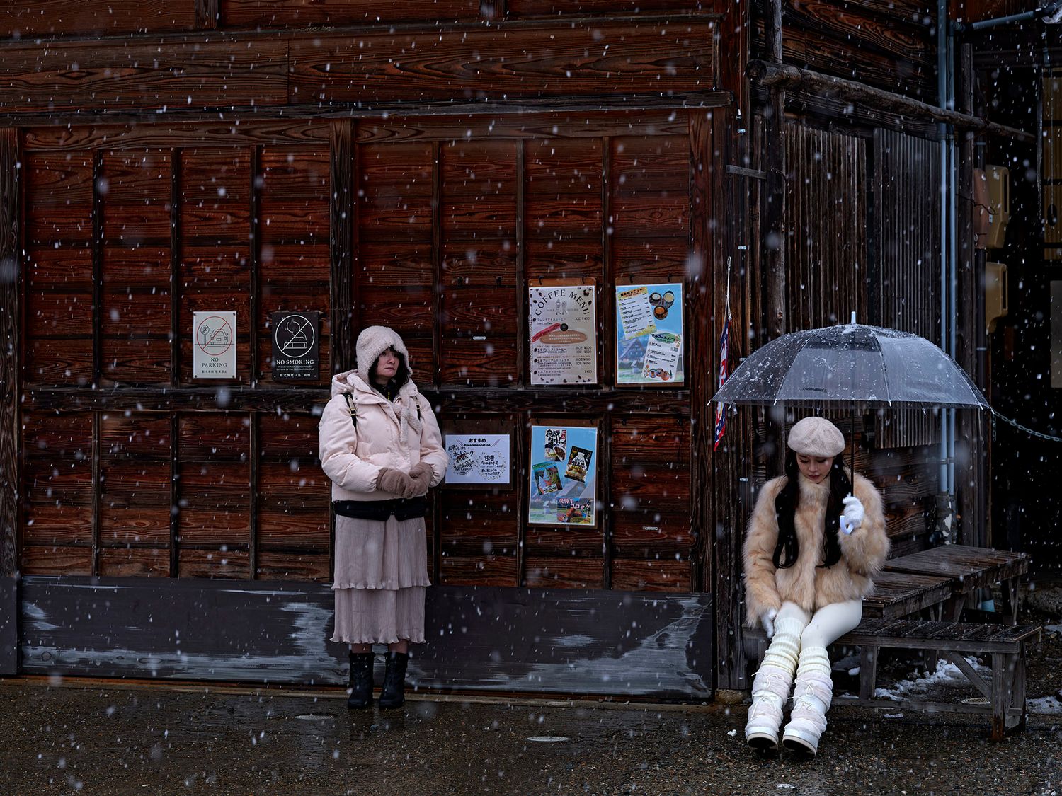 Two women wait outside in snowfall, one holding an umbrella, both wearing winter coats near a wooden building.