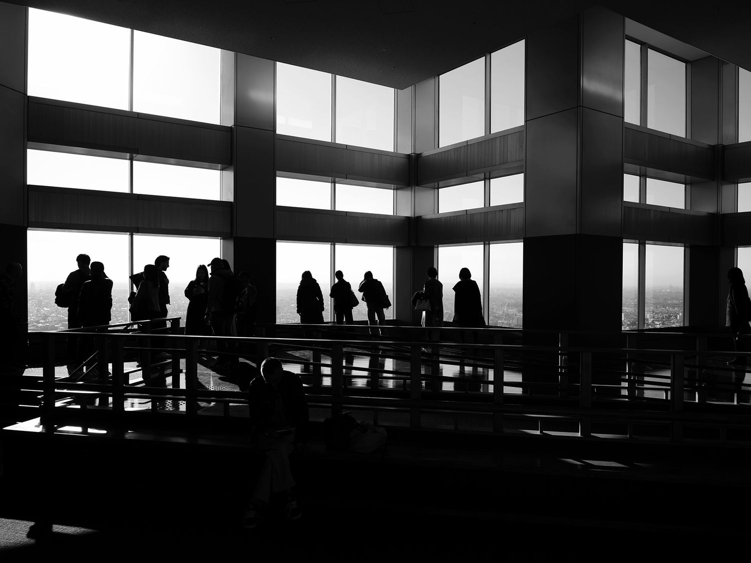 Silhouettes of people standing near large windows in a modern building, captured in dramatic black and white photography.