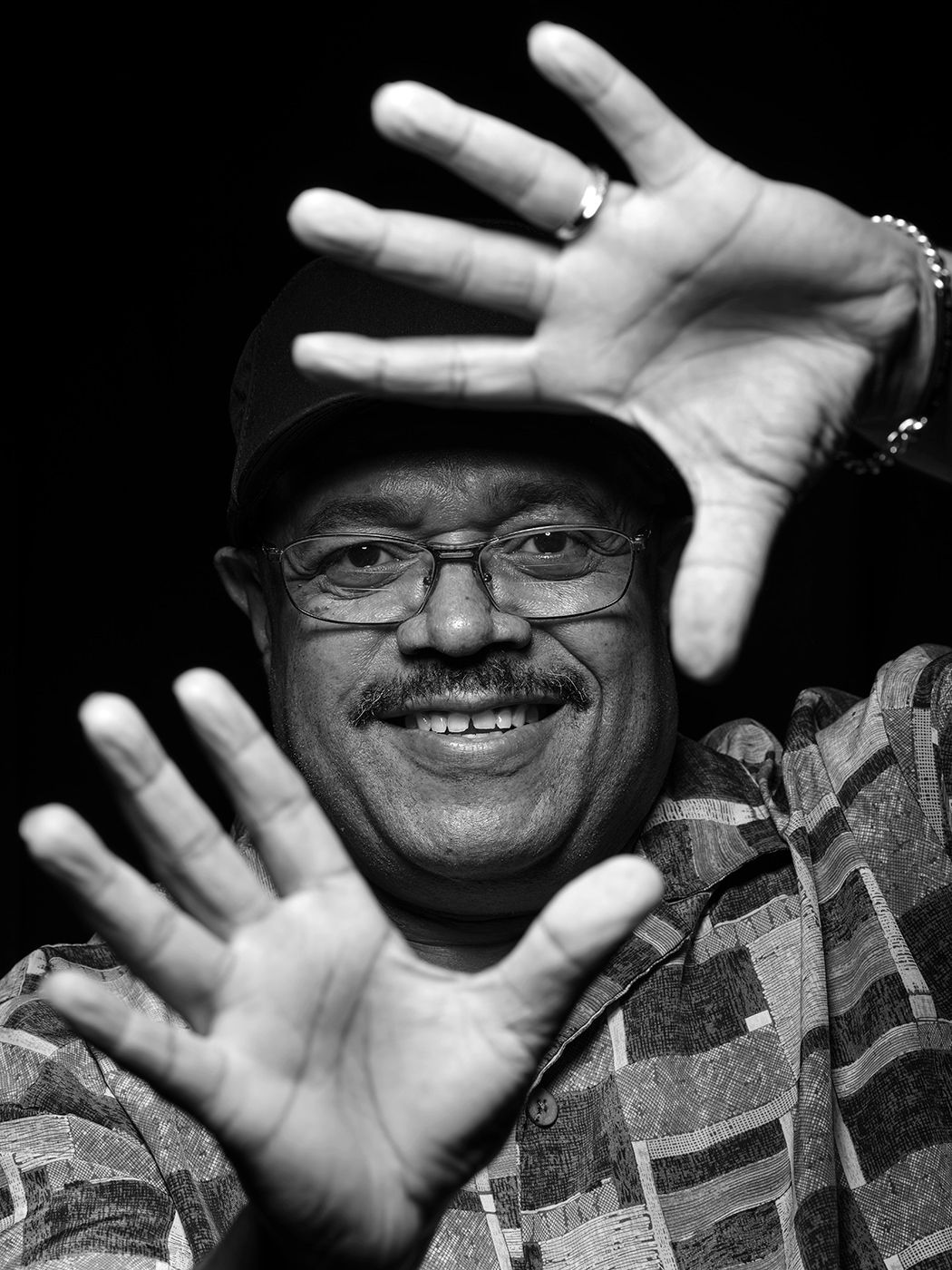 Smiling man with glasses raises both hands toward camera in a black and white portrait photo. Dennis Chambers