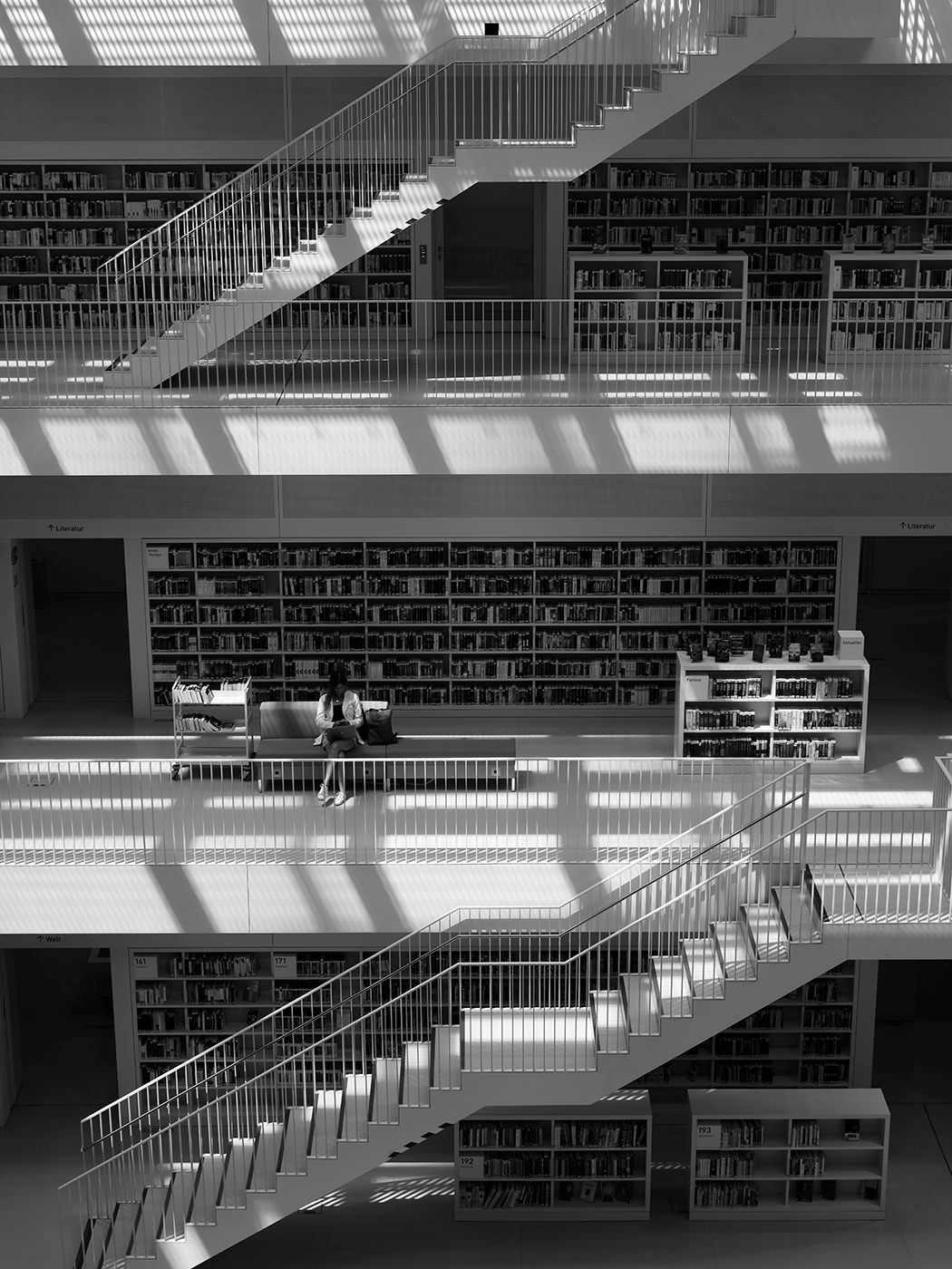 Black and white photo of a modern multi-level library interior with staircases, bookshelves, and a lone reader.