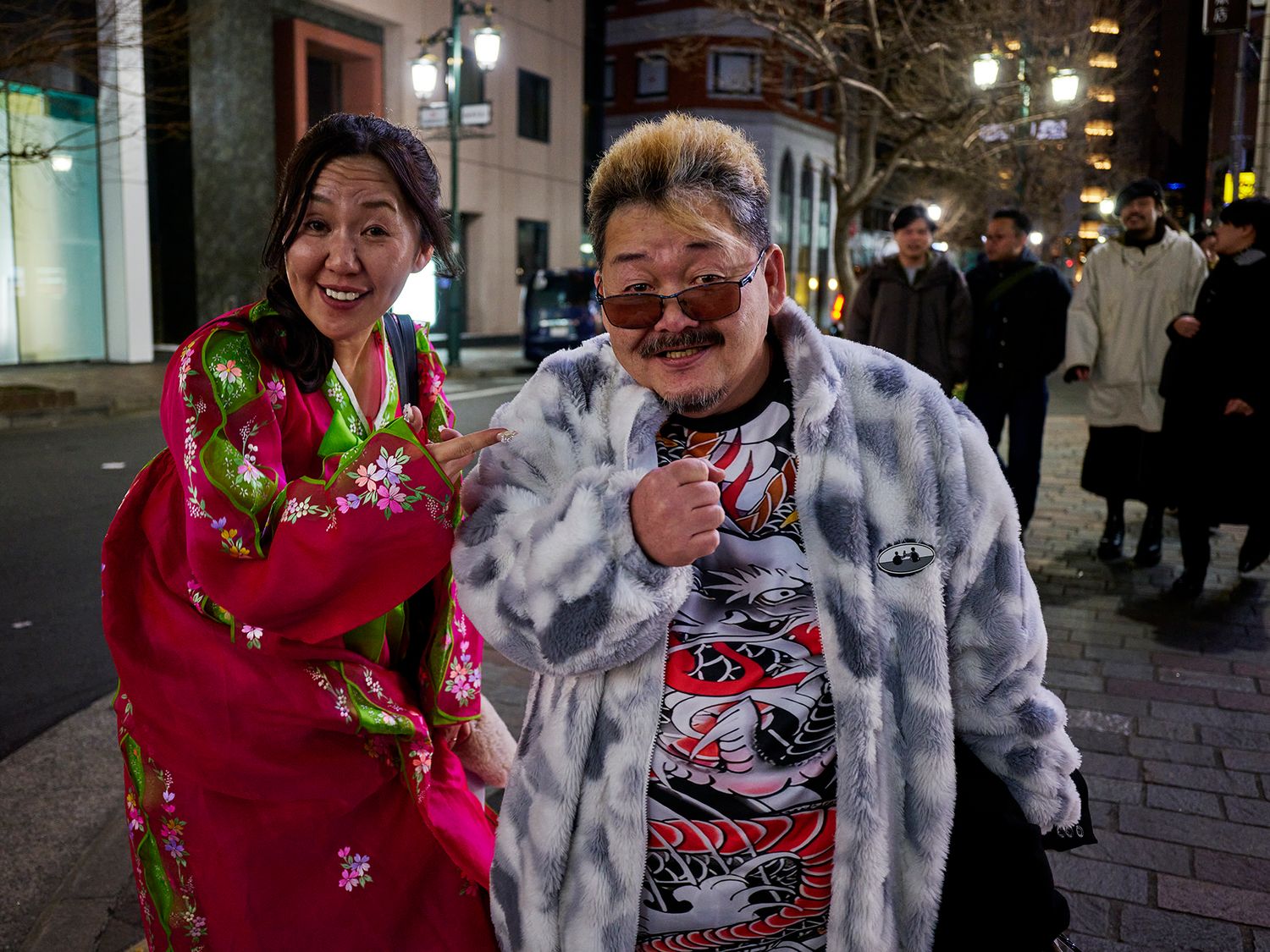 Two Asian women smiling outdoors at night, one in pink floral kimono, other in fur coat with red sunglasses.