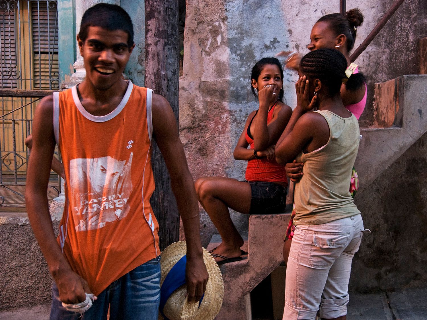 Young people socializing on a colorful urban street corner in Cuba, with a man in an orange tank top in the foreground.