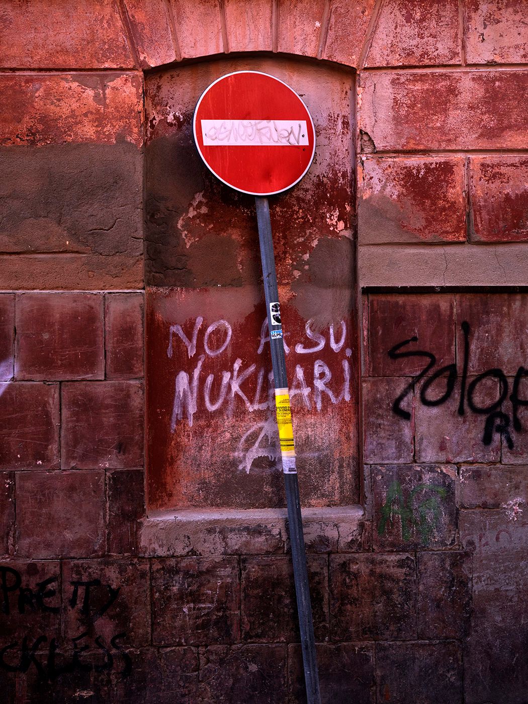 Red no-entry sign on a pole against a graffiti-covered brick wall with anti-nuclear text in urban setting.