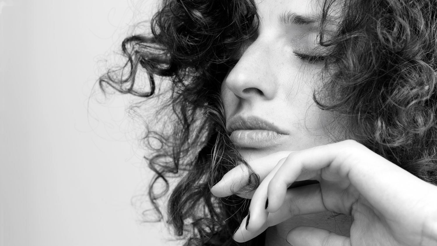 Black and white close-up portrait of a curly-haired woman resting her chin on her hands thoughtfully.