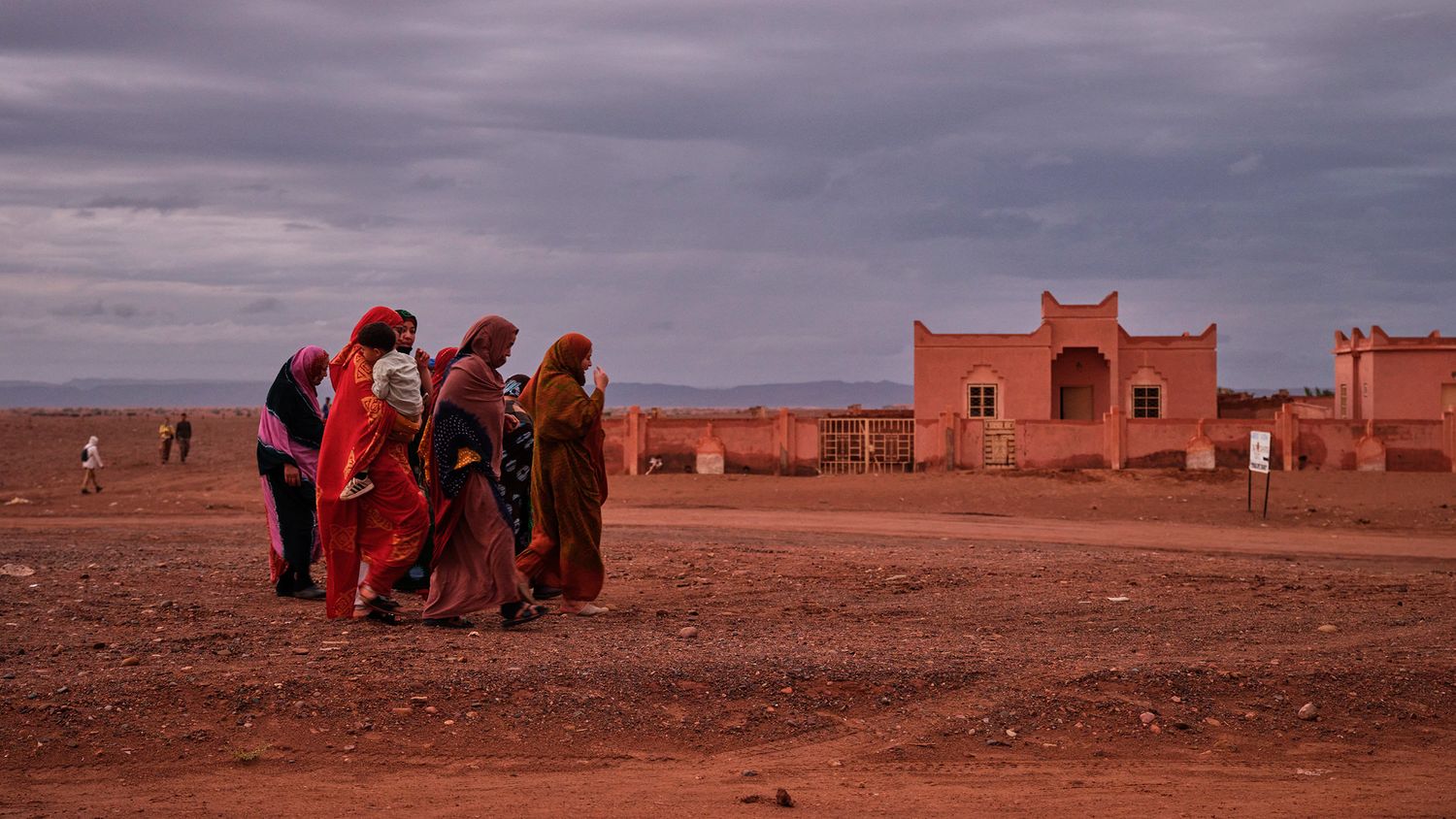 Group of colorfully dressed women walking across a red dirt landscape near a terracotta building at dusk.
