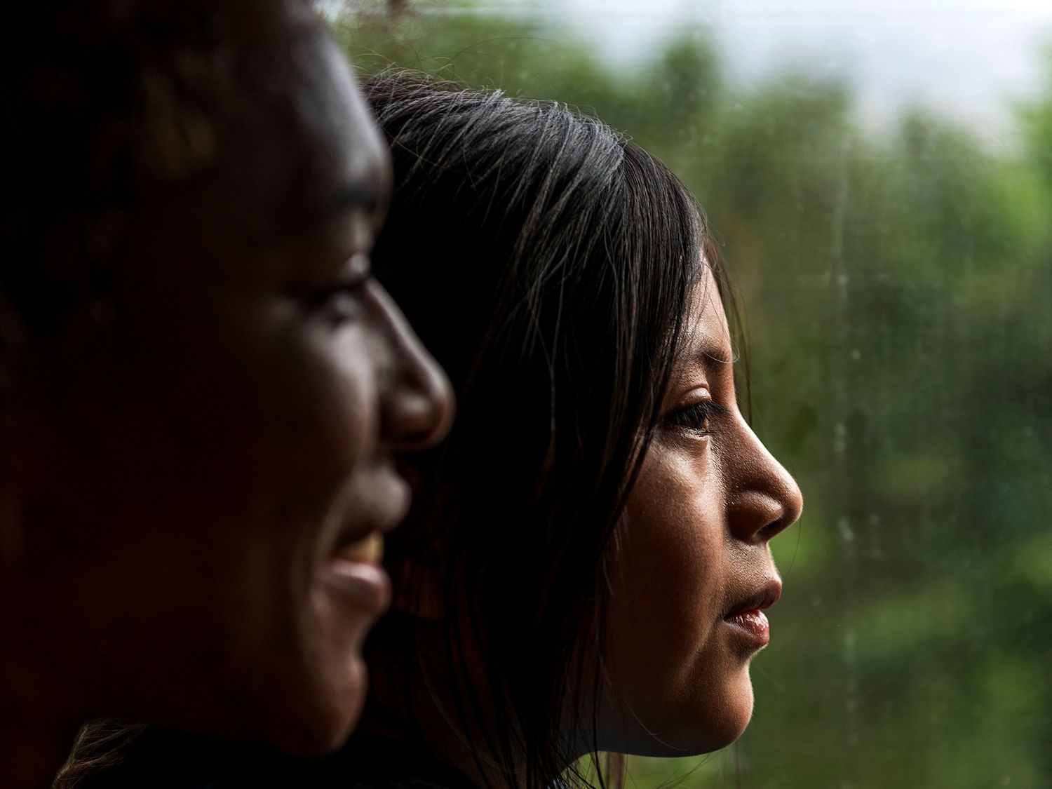 Two women in profile looking out a rain-streaked window in soft natural light.