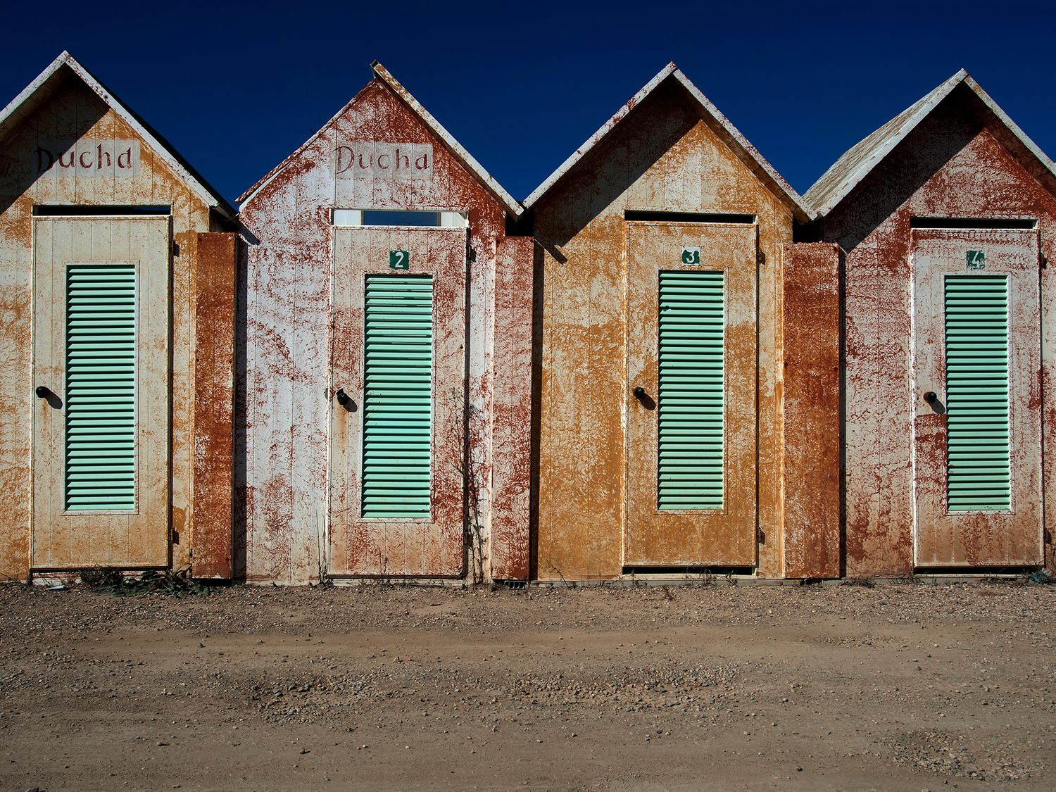 Row of weathered wooden beach huts with green shuttered doors on sandy ground under a clear blue sky.