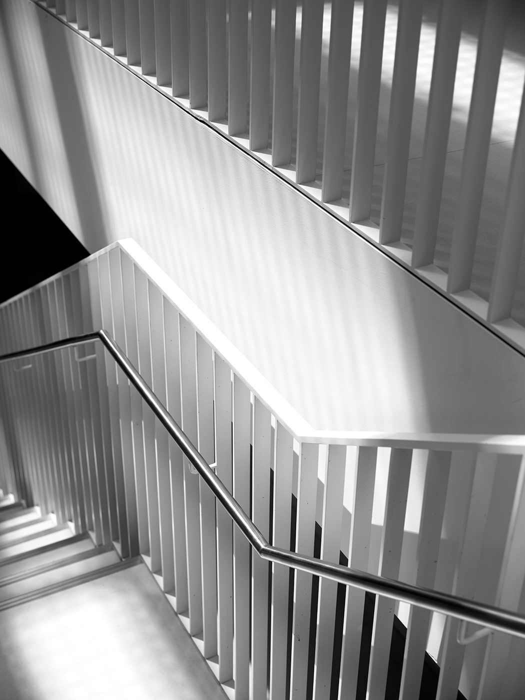 Black and white architectural photo of modern staircase with white balusters and handrail, geometric lines and light contrast.