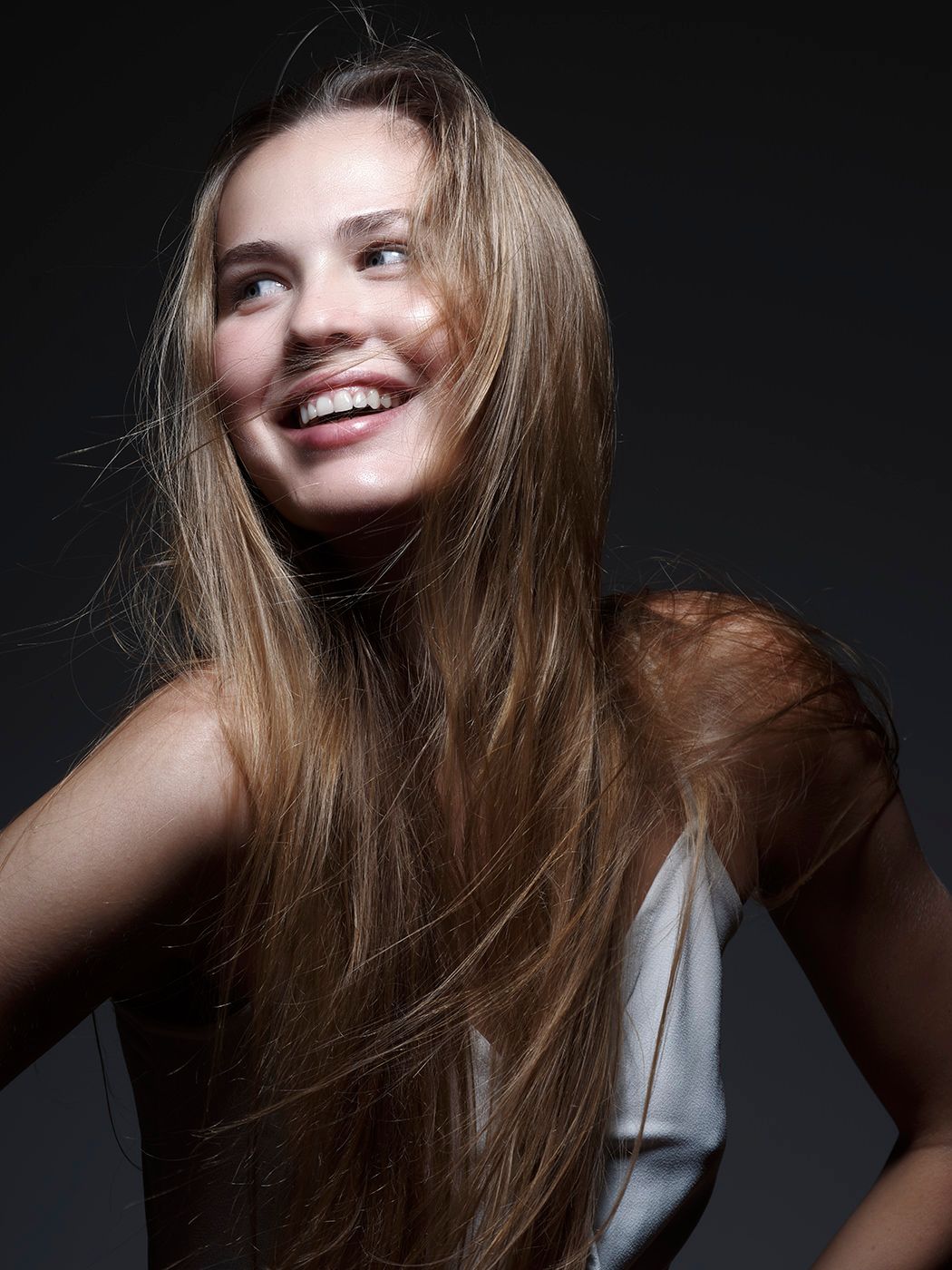 Young woman with long blonde hair smiling joyfully against a dark background in a white top.