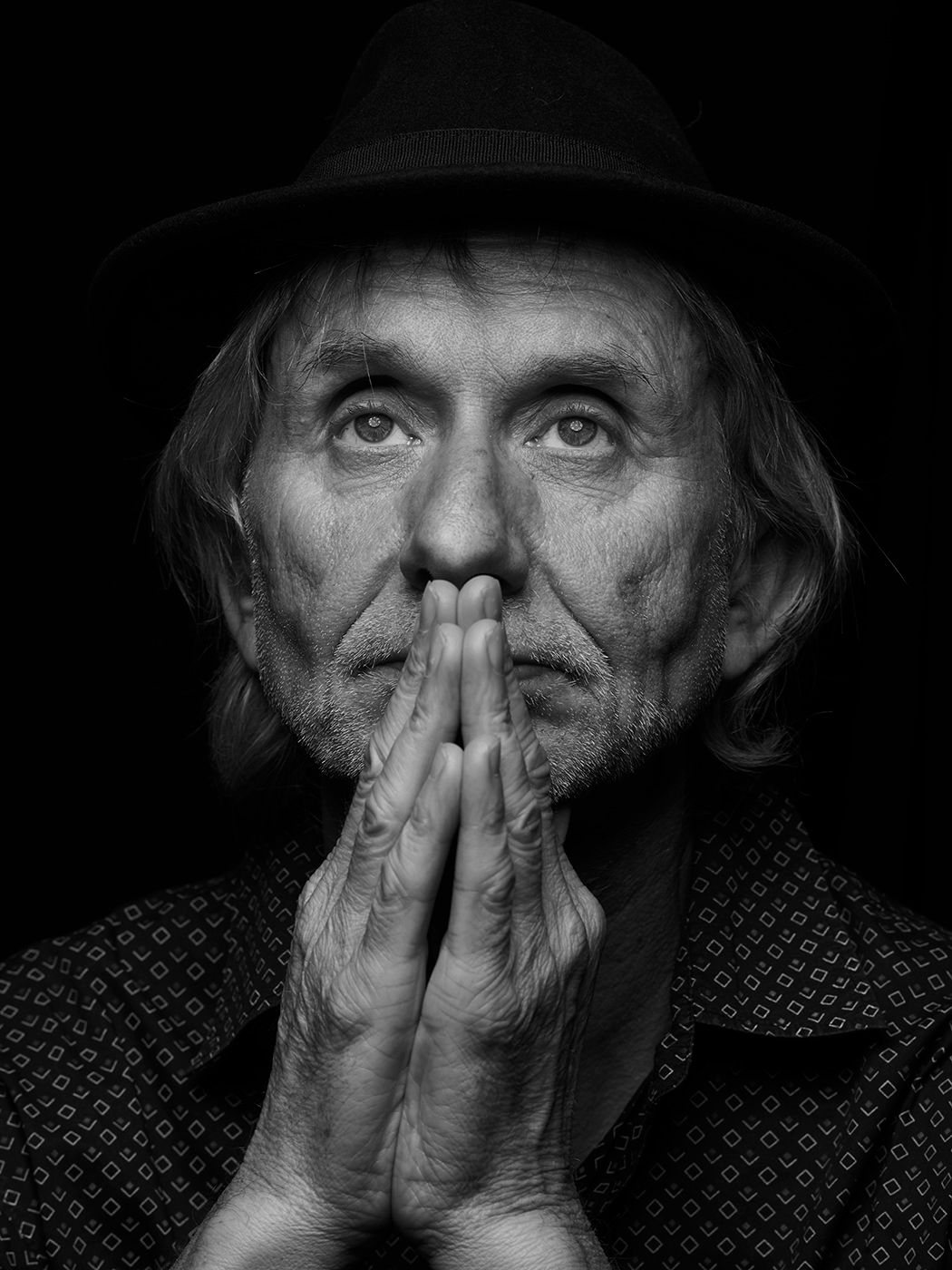Elderly man in black hat with hands clasped in prayer pose, dramatic black and white portrait photography. Erik Truffaz