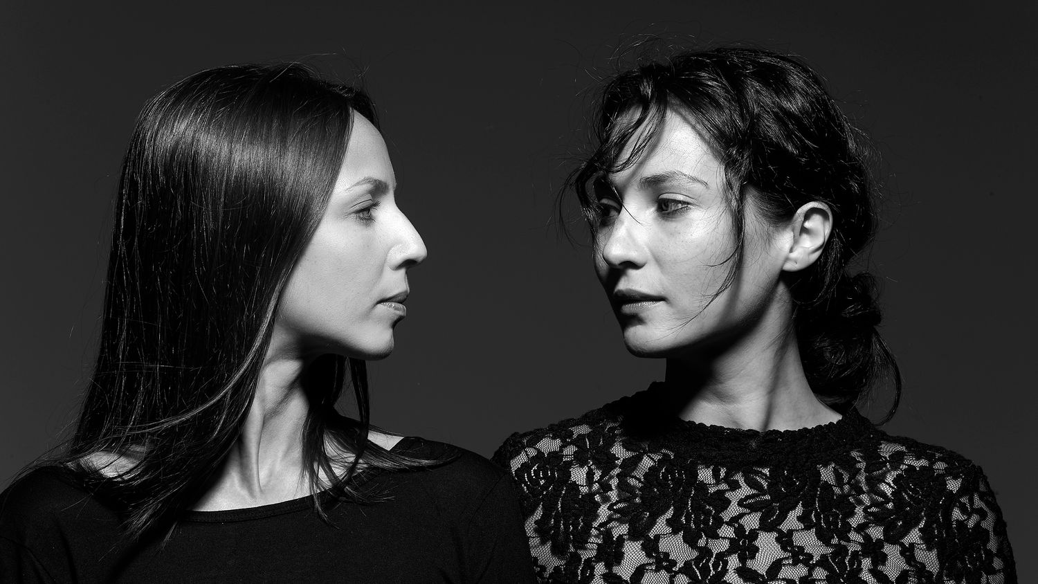 Two women in black clothing face each other in a dramatic black and white portrait photo.