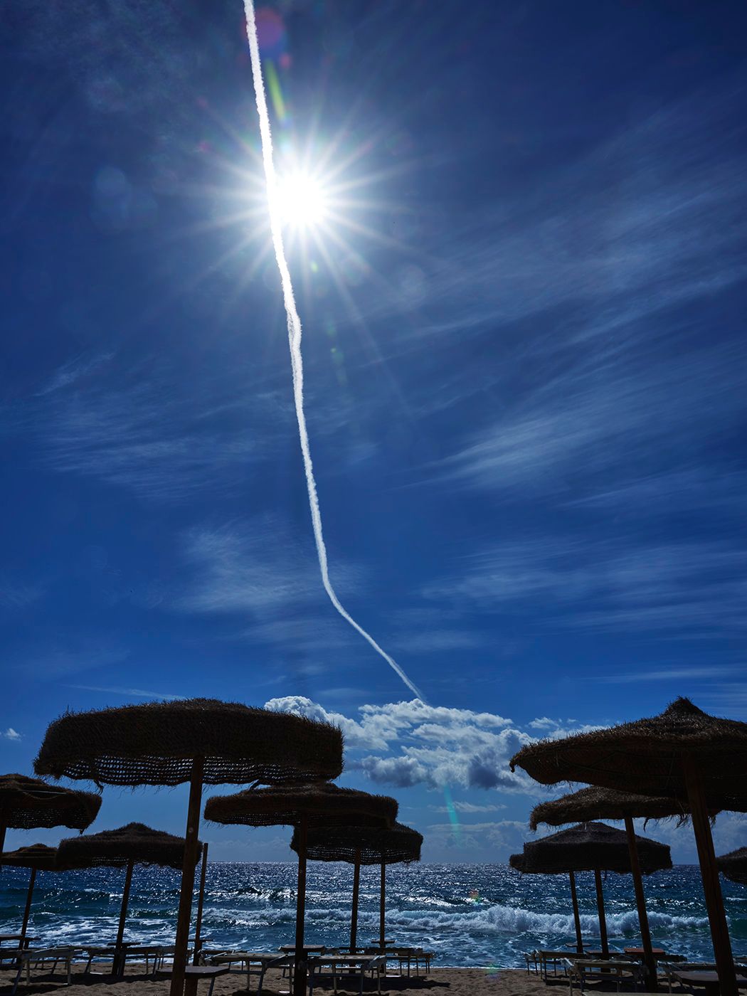 Beach umbrellas under bright sun with blue sky, lens flare, and ocean view on a sunny day.