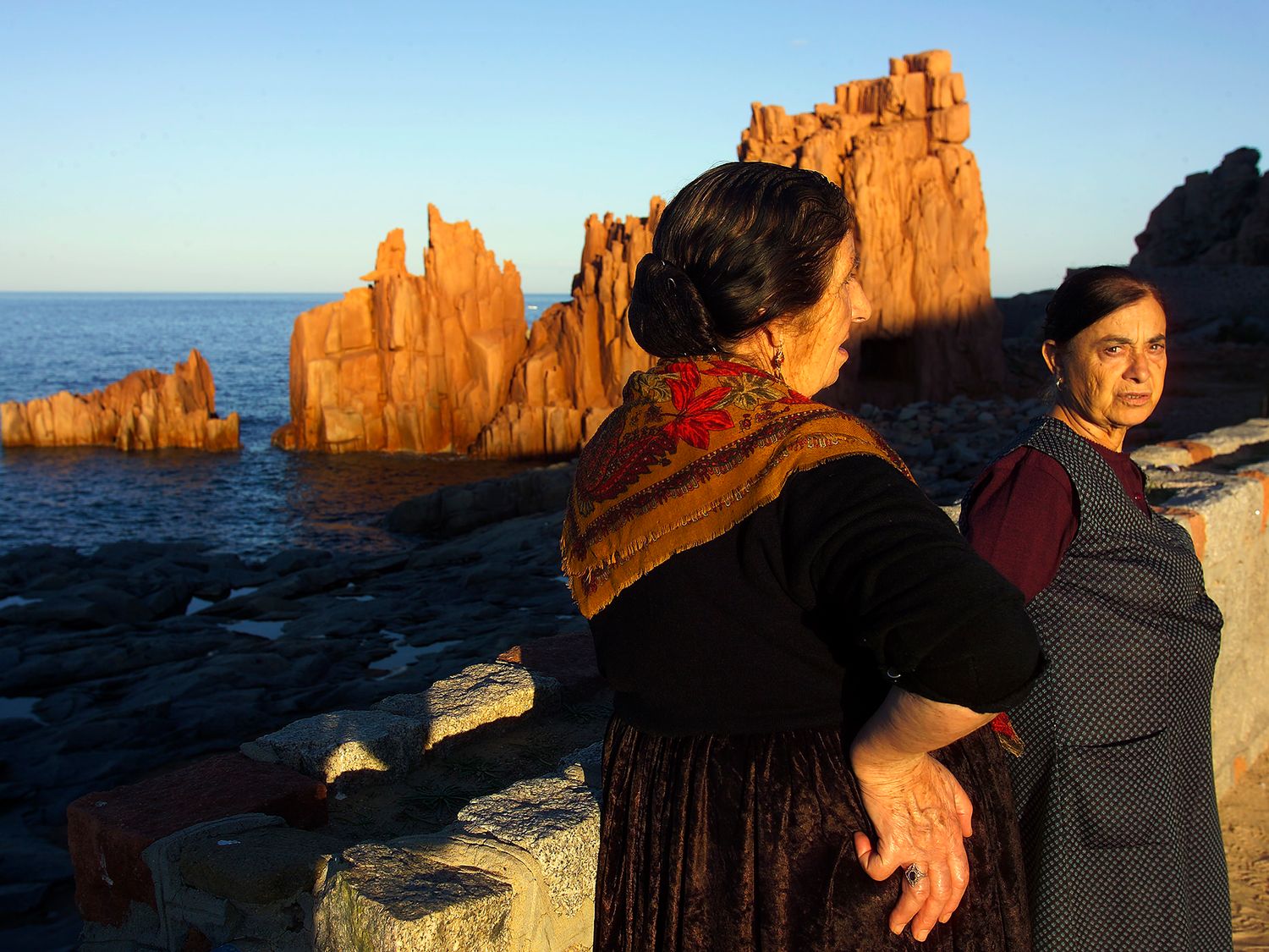 Two women in traditional dress stand on rocky shore with dramatic red rock formations and blue sea at sunset.