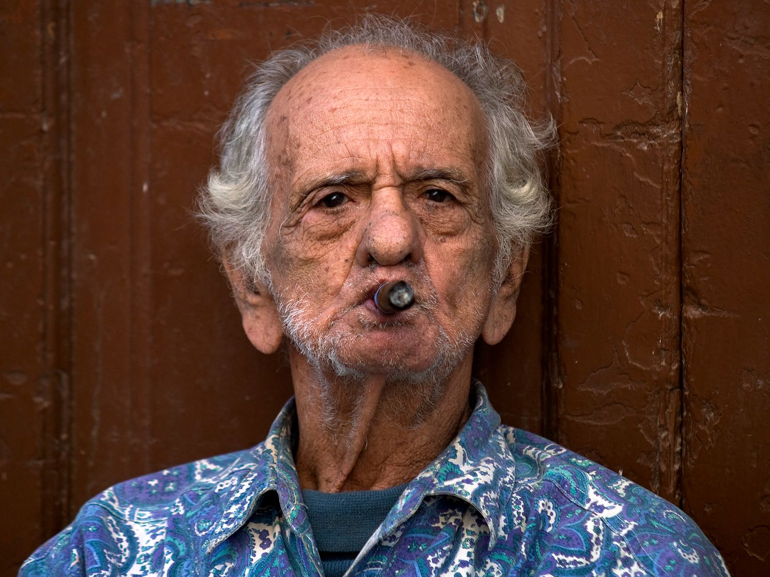 Elderly man with gray hair smoking a cigar, wearing a blue floral shirt, posing against a wooden wall.