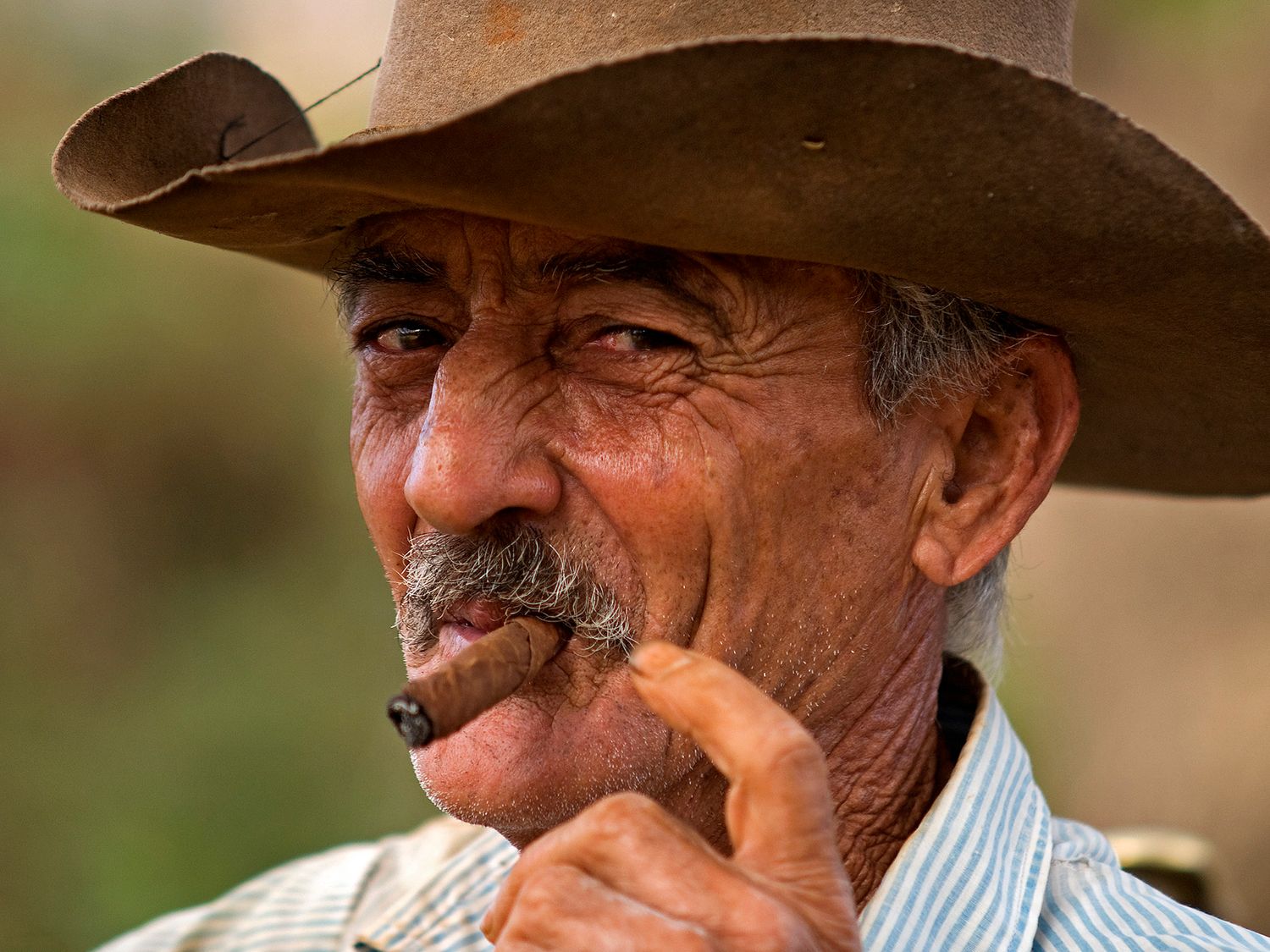 Elderly man wearing a brown cowboy hat smoking a cigar, with a mustache and weathered face.