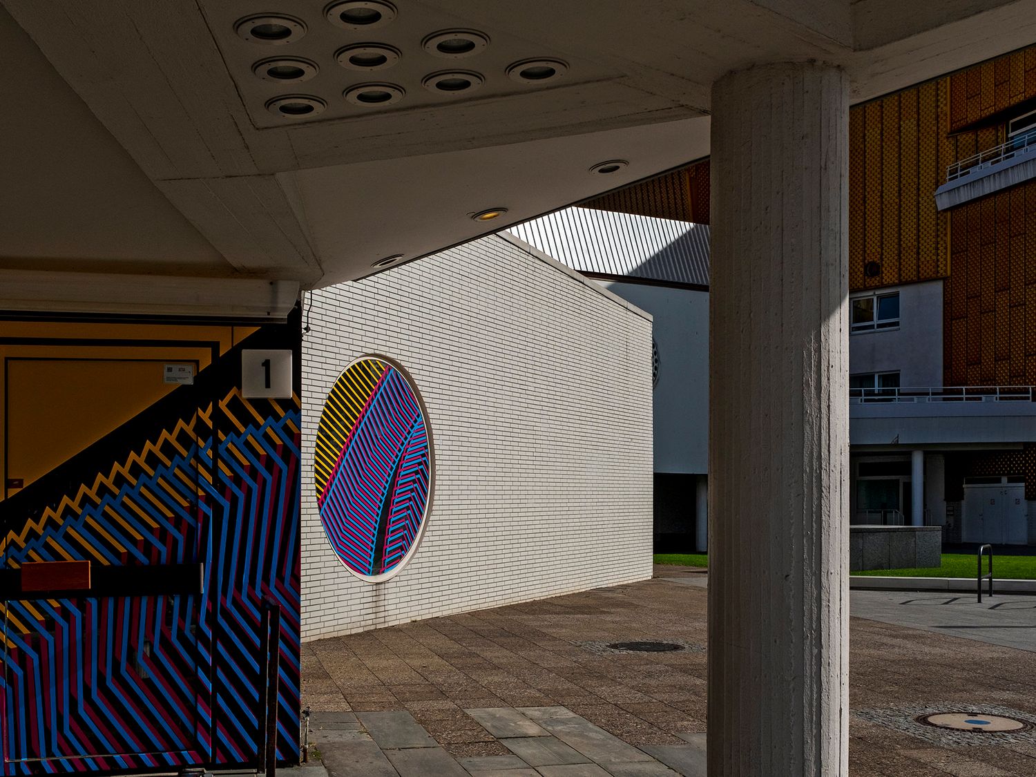 Colorful op-art mural on a white building facade, viewed through concrete columns in an urban plaza.