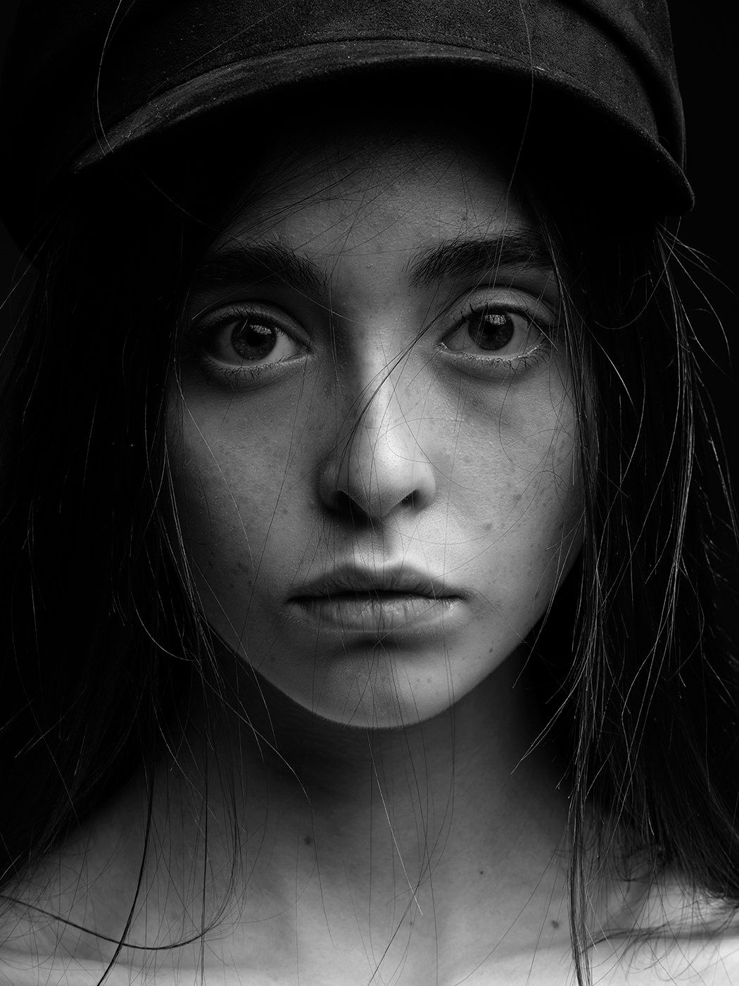 Black and white portrait of a young woman with freckles wearing a cap, intense gaze, dark moody background.