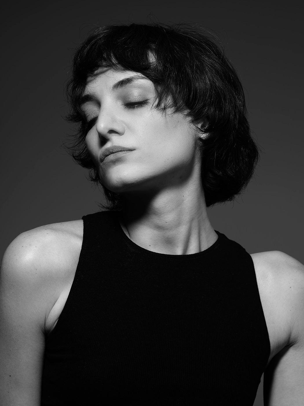 Black and white portrait of a young woman with short wavy hair wearing a black sleeveless top, eyes closed.