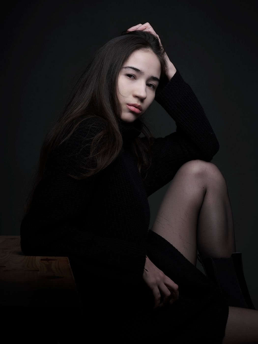 Young woman in black dress and tights poses elegantly against dark studio background with hand in hair.