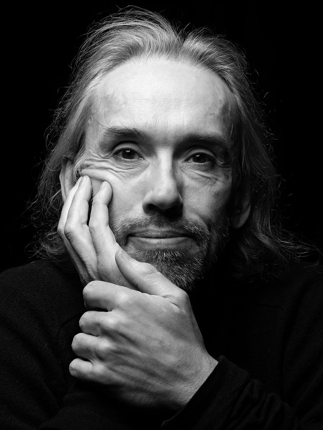 Black and white portrait of a middle-aged man with long hair resting his chin on his hand against a dark background. Björn Meyer