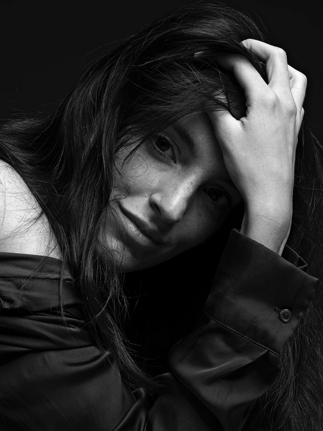 Black and white portrait of a woman with long dark hair, hand raised to her head, wearing a dark jacket.