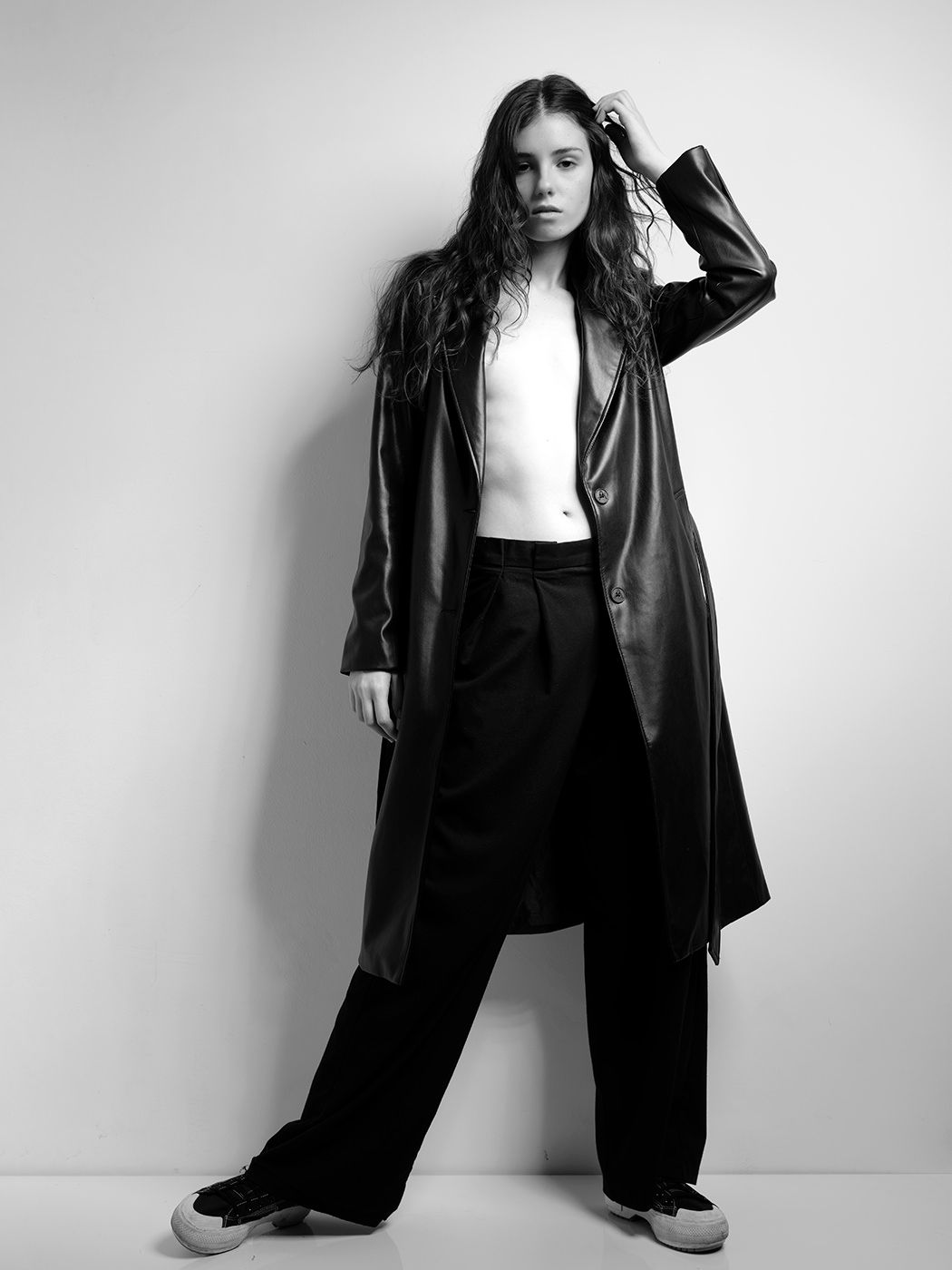 Black and white photo of a woman in a long leather coat, wide-leg pants, and white top posing against a wall.