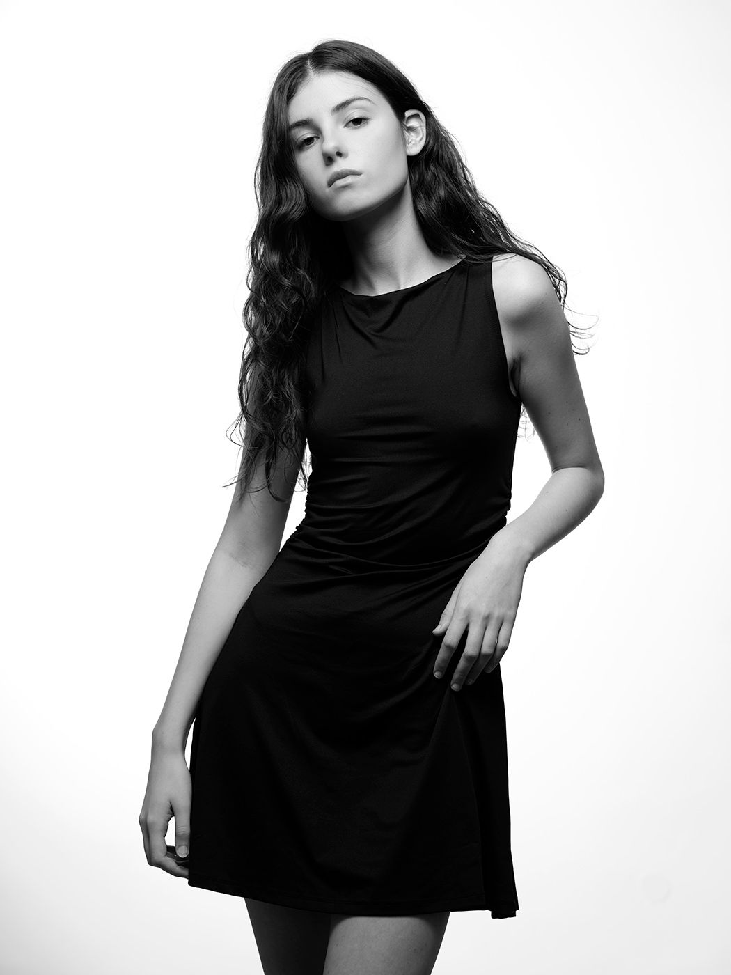Black and white photo of a young woman in a sleeveless black mini dress posing against a white background.